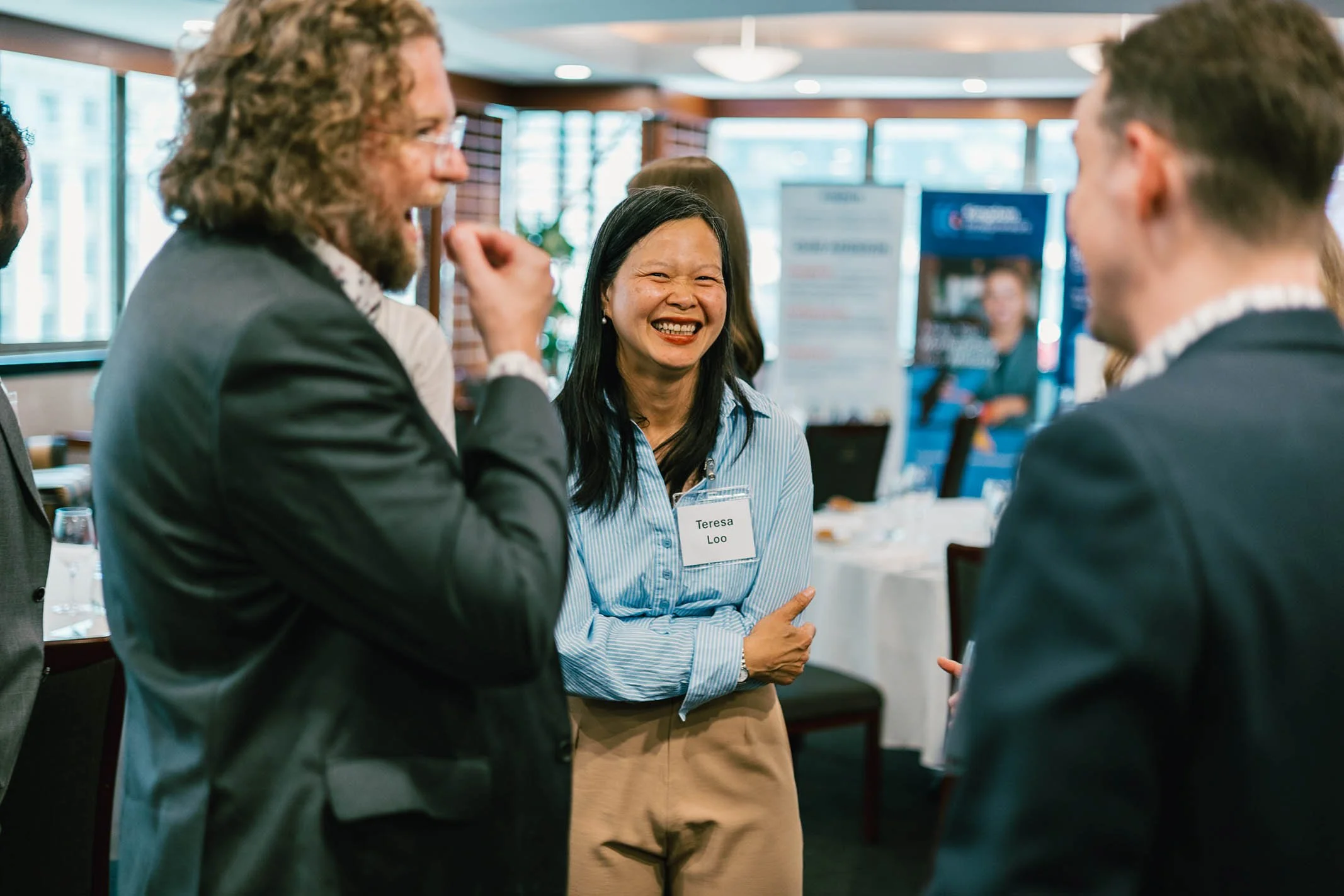 Multiple people at a networking event engaging in conversation, with one woman smiling and wearing a name tag that says Teresa Loo, and others in business attire.