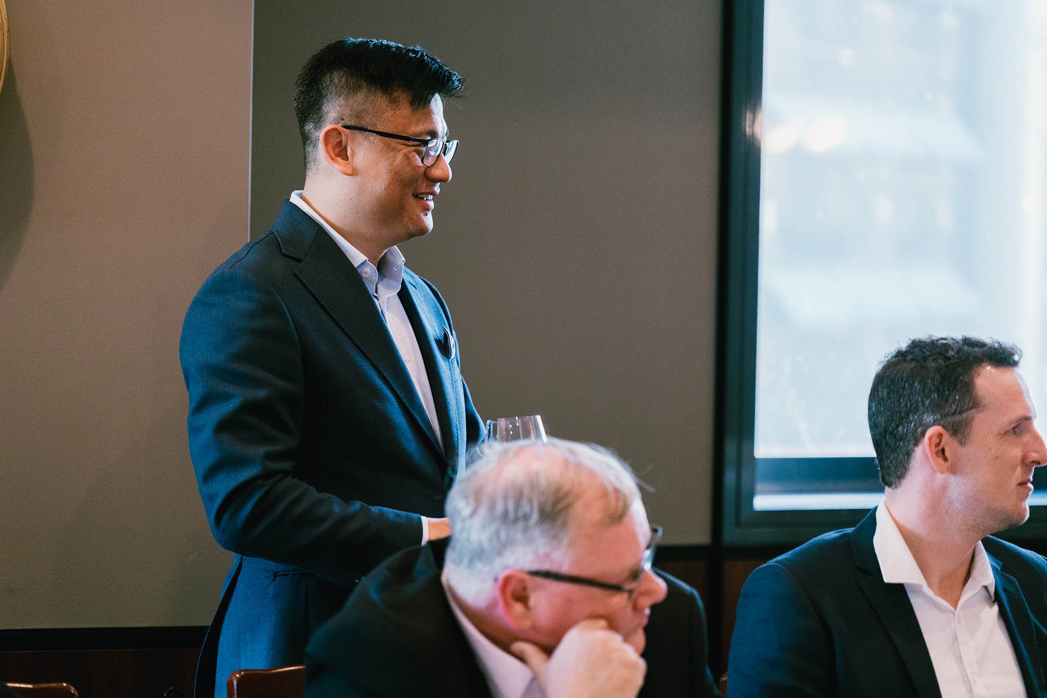 A man in a dark suit and glasses standing with a glass of wine during a business meeting or conference, three other men seated at the table, one with glasses and gray hair, the other with dark hair, in a room with large window and natural light.