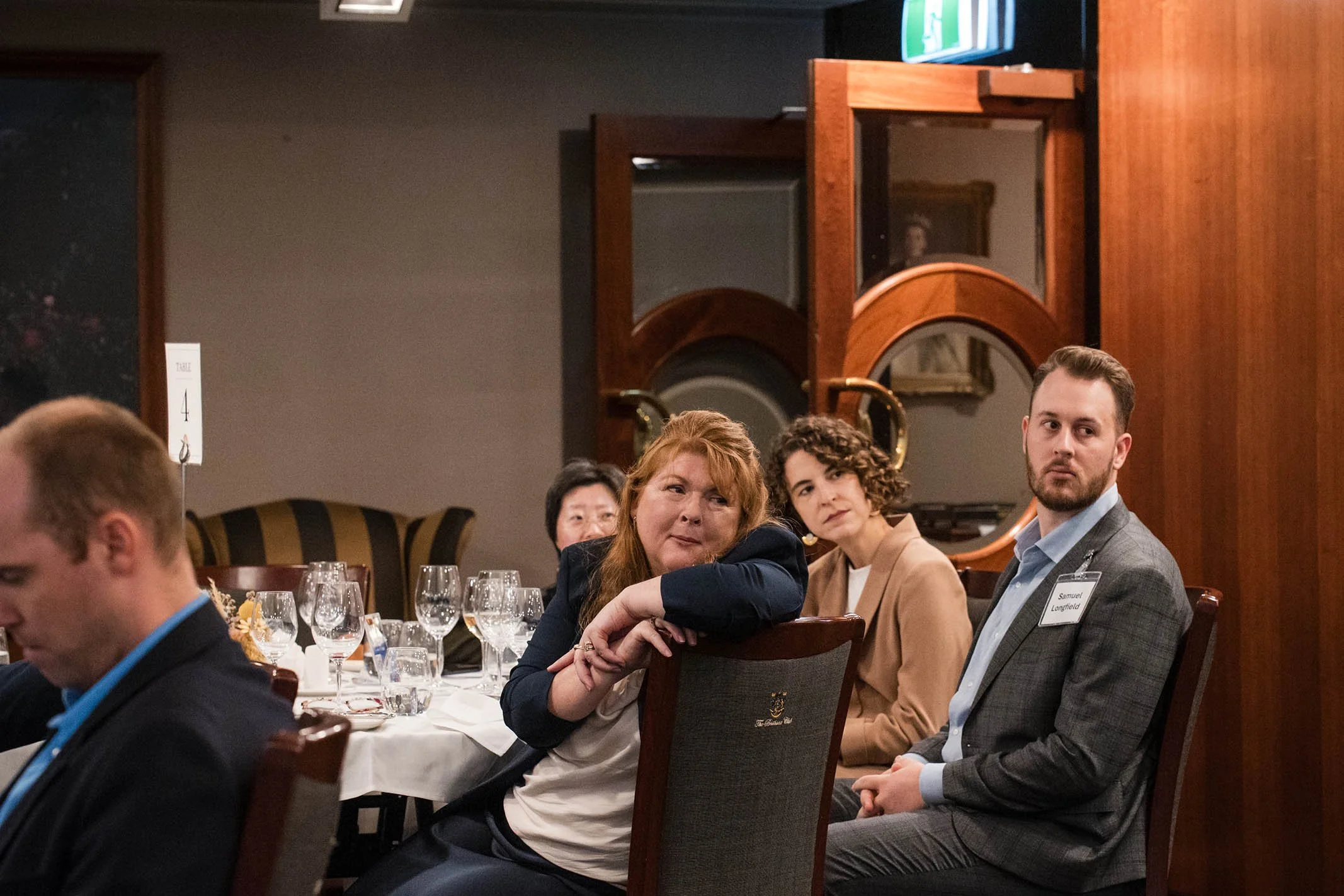 People seated at a banquet table during a formal event, with wine glasses and table settings, in a room with wood decor and a TV on the wall.