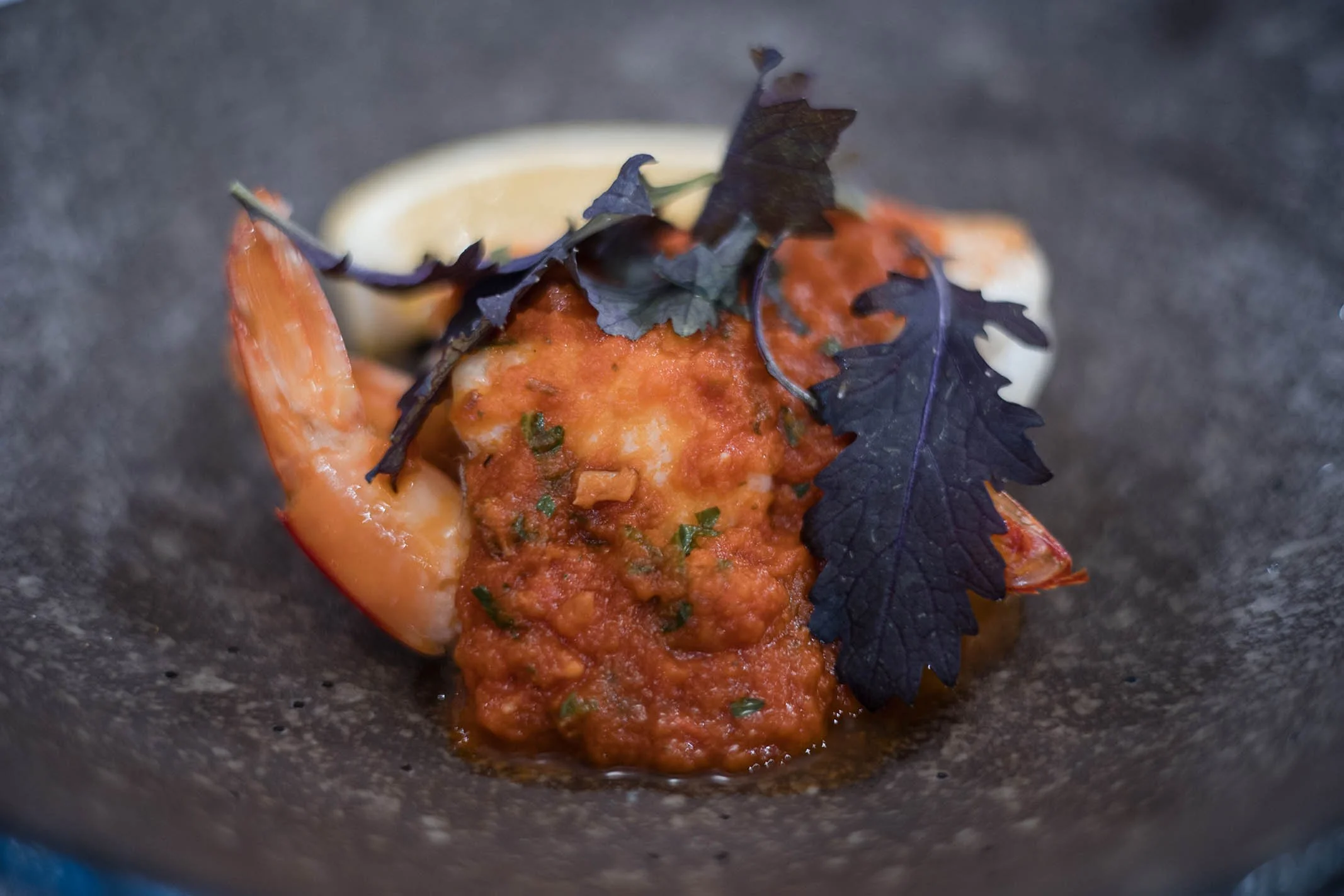 Close-up of a plated seafood dish featuring a cooked shrimp, a lemon wedge, dark purple herb leaves, and a tomato-based sauce with herbs.