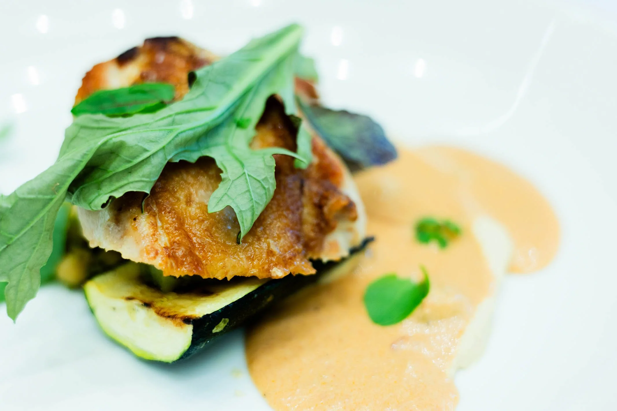 A plated gourmet dish featuring a grilled vegetable, leafy greens on top, and a creamy orange sauce on the side.
