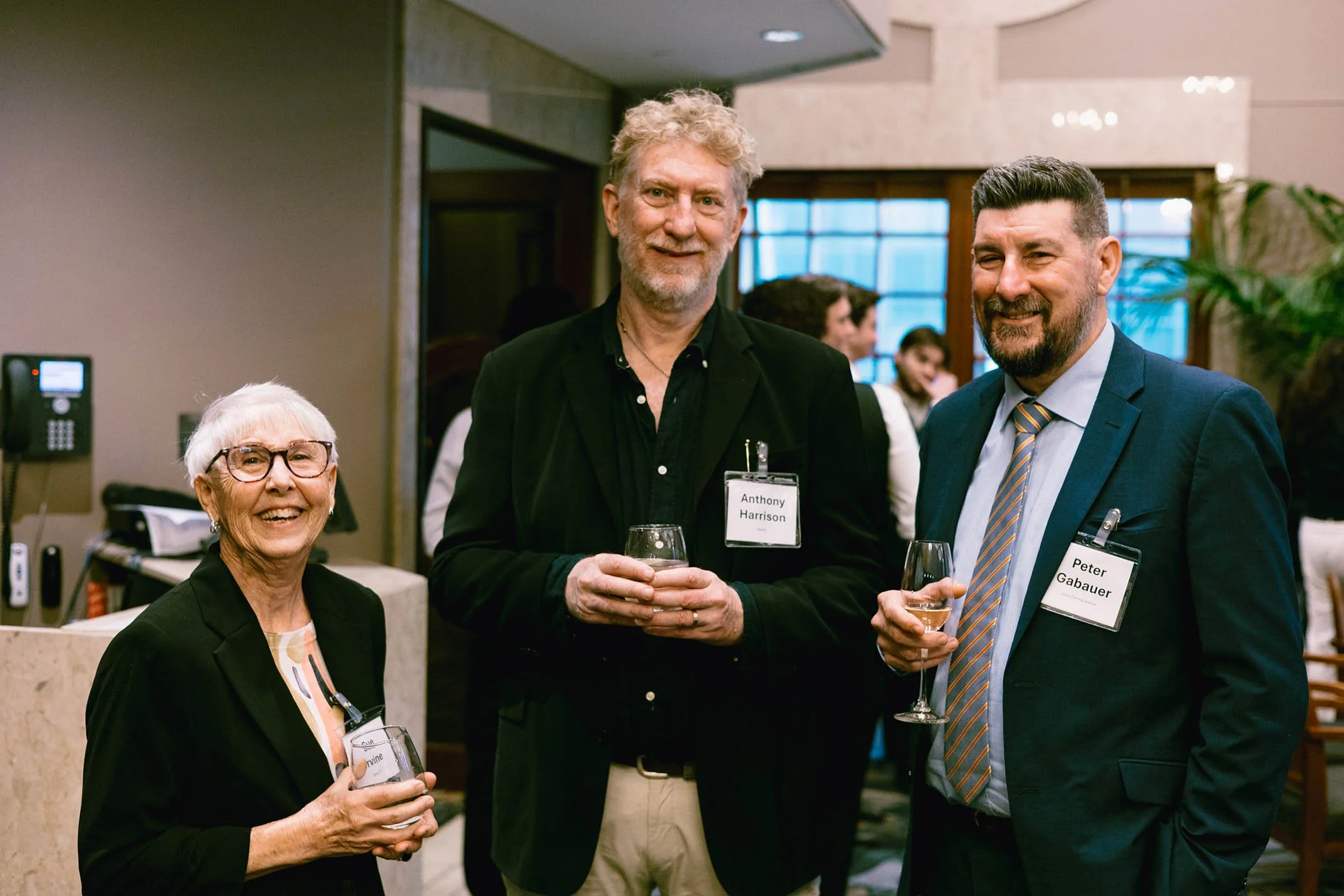 Three people at a social event, smiling and holding drinks. An elderly woman with glasses and a black blazer, a tall man with curly blonde hair in a black blazer, and a man with short dark hair and a beard in a blue suit and striped tie.