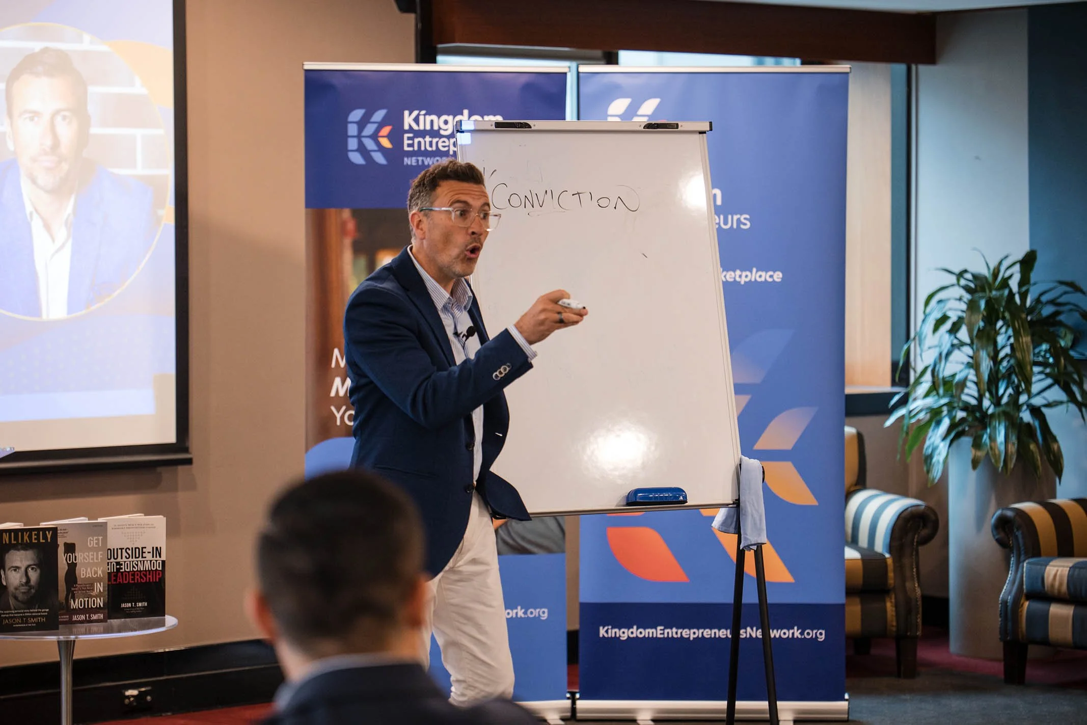 A man in a blue blazer and glasses is giving a presentation about conviction at a conference, standing beside a whiteboard with the word 'Conviction' written on it. There are banners for Kingdom Entrepreneurs Network behind him and books on a table t