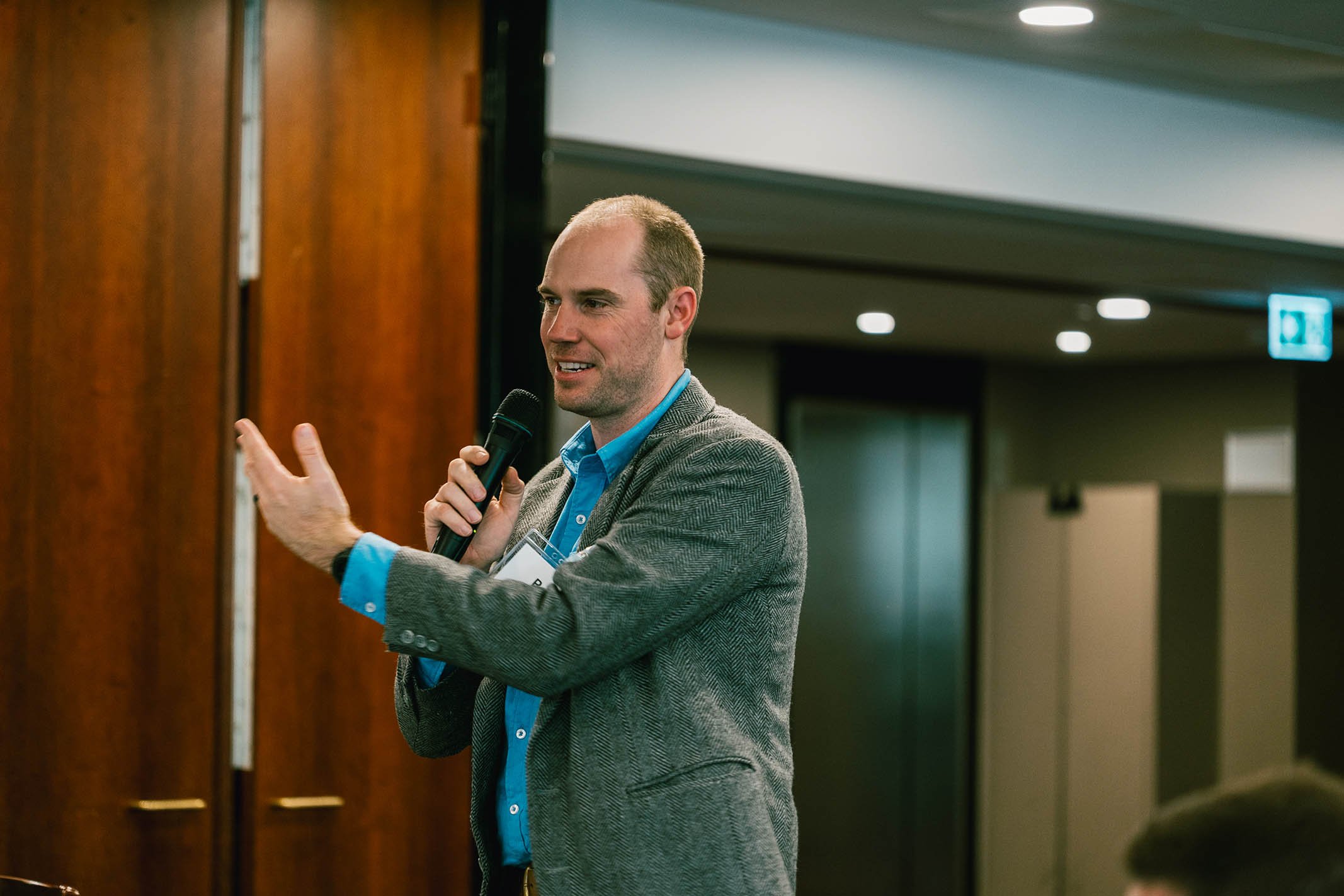 A man in a gray blazer and bright blue shirt speaking into a microphone during a presentation or speech in an indoor conference room.