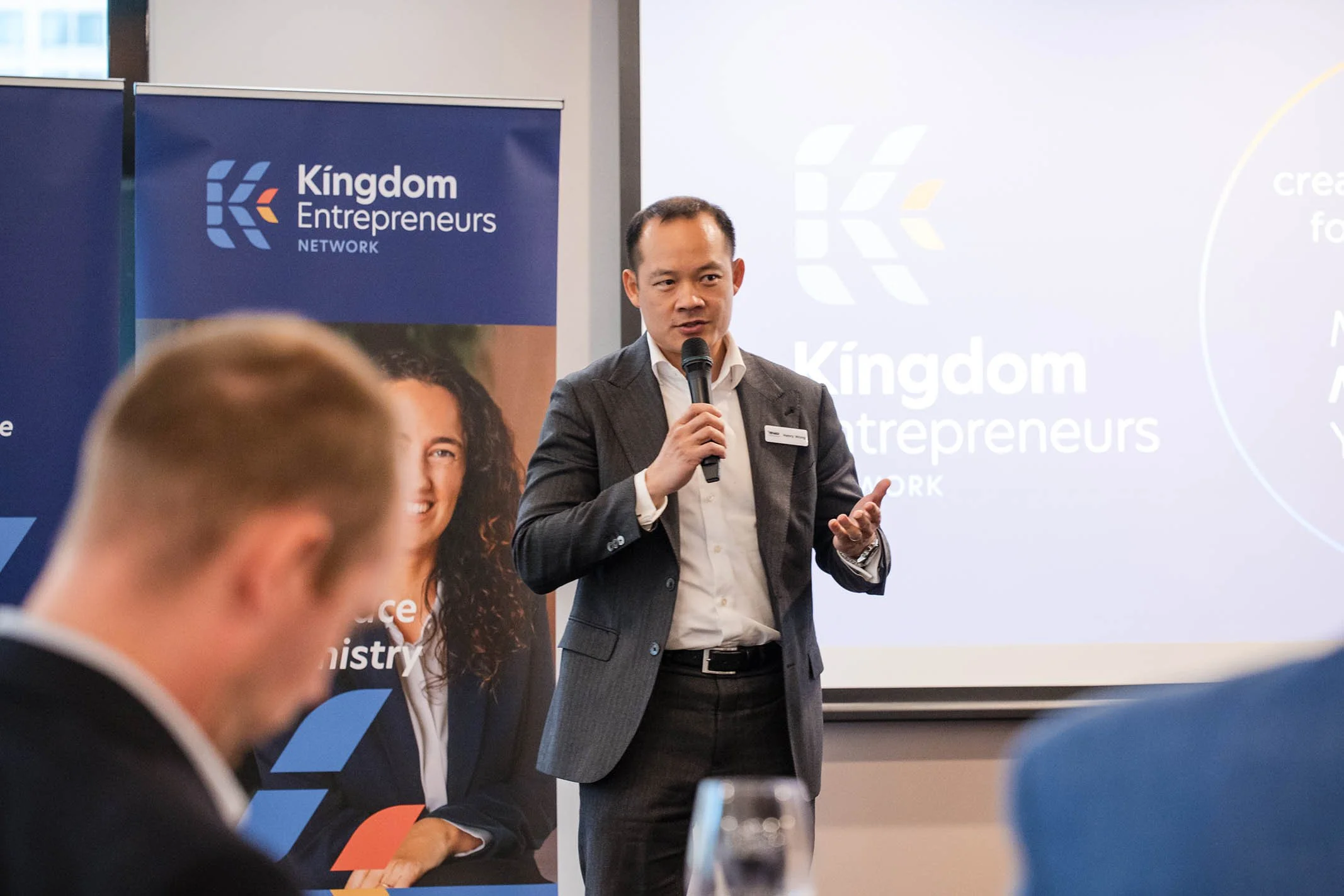 A man in a gray suit holding a microphone giving a presentation at a Kingdom Entrepreneurs Network event, with a large screen displaying the network's logo and a blue banner with a woman's face and logo in the background.