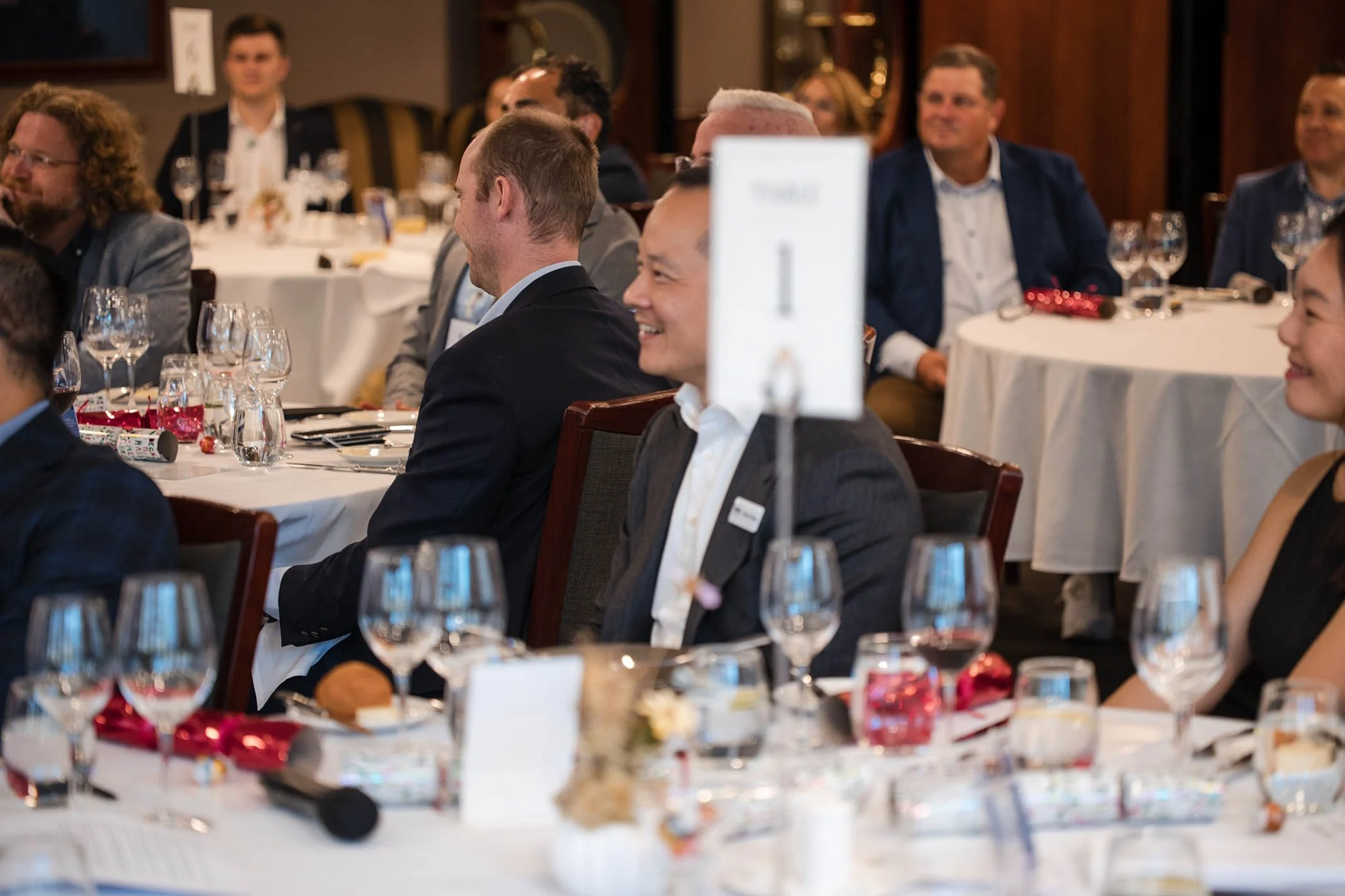 People sitting at round tables with white tablecloths, glasses, and decorations, attending an event or banquet in a formal setting, with some individuals smiling and engaging.