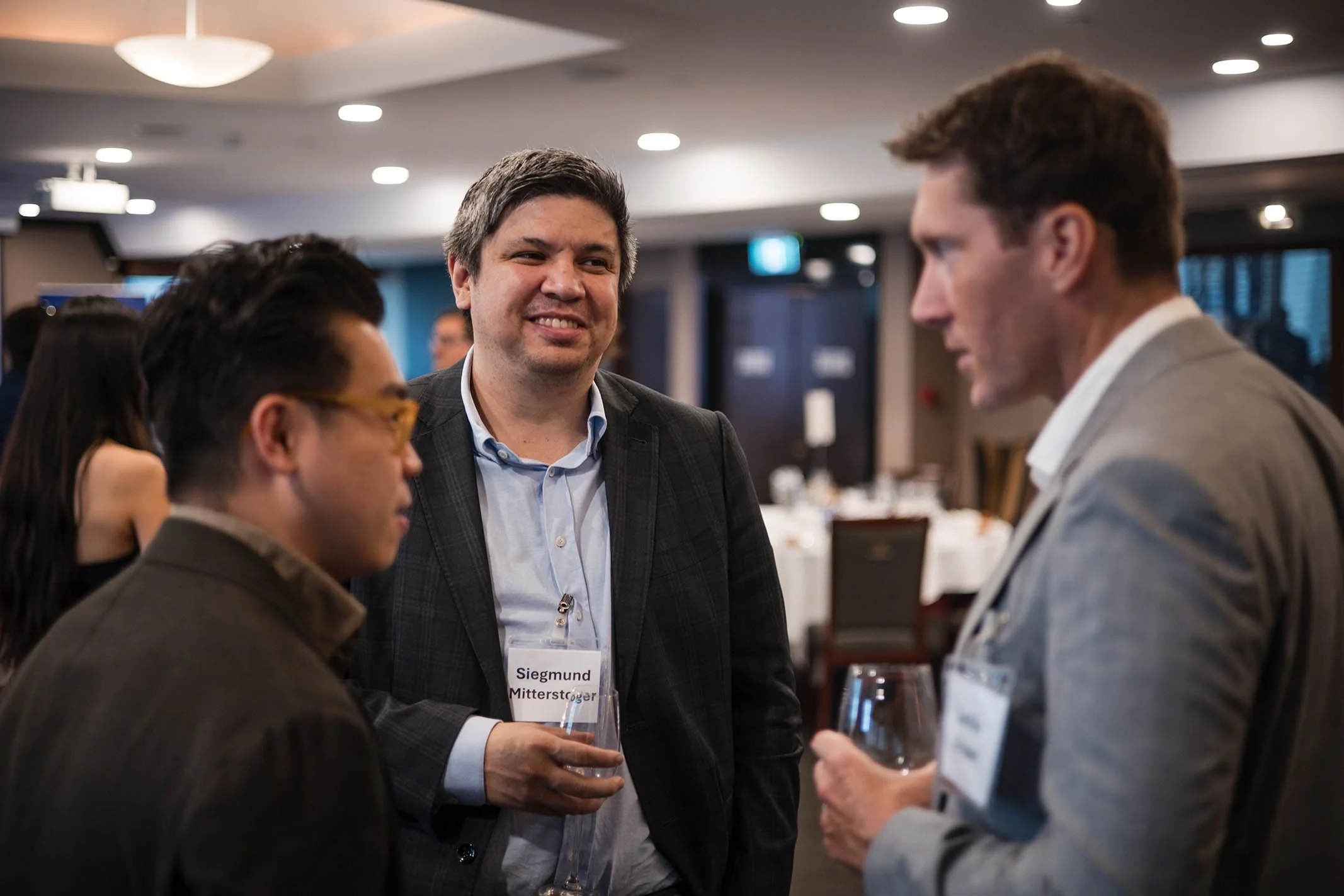 Three men engaged in conversation at a professional networking event, with tables and attendees in the background.