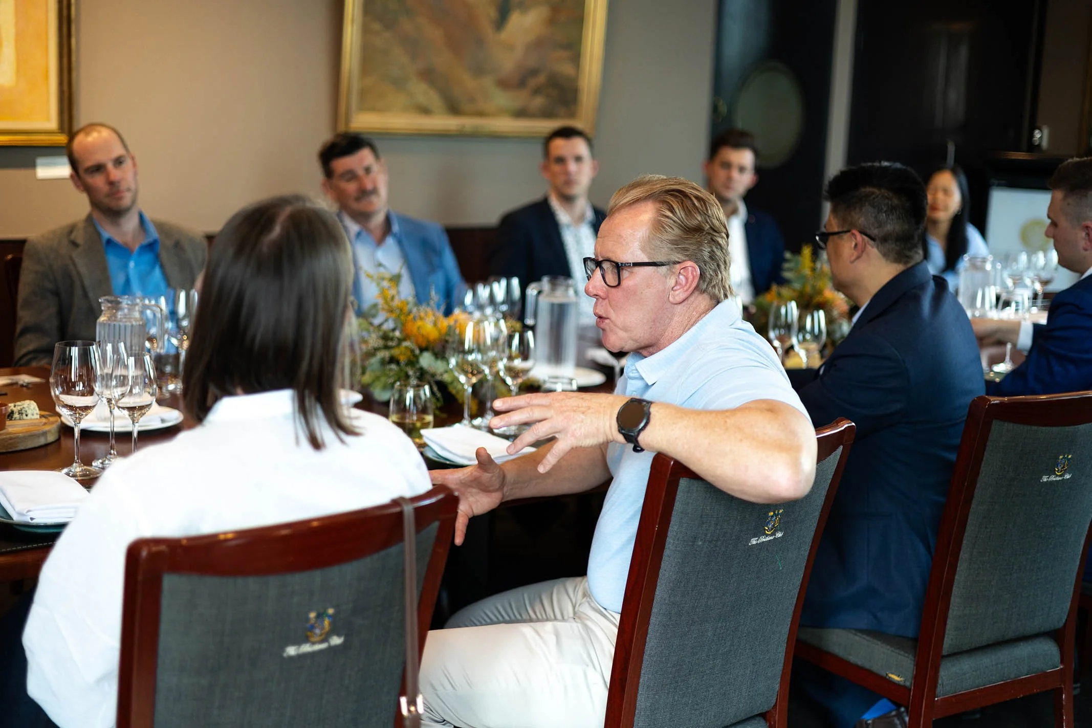 A group of professionally dressed people seated at a rectangular dining table engaged in conversation, with greenery and paintings on the wall behind them.