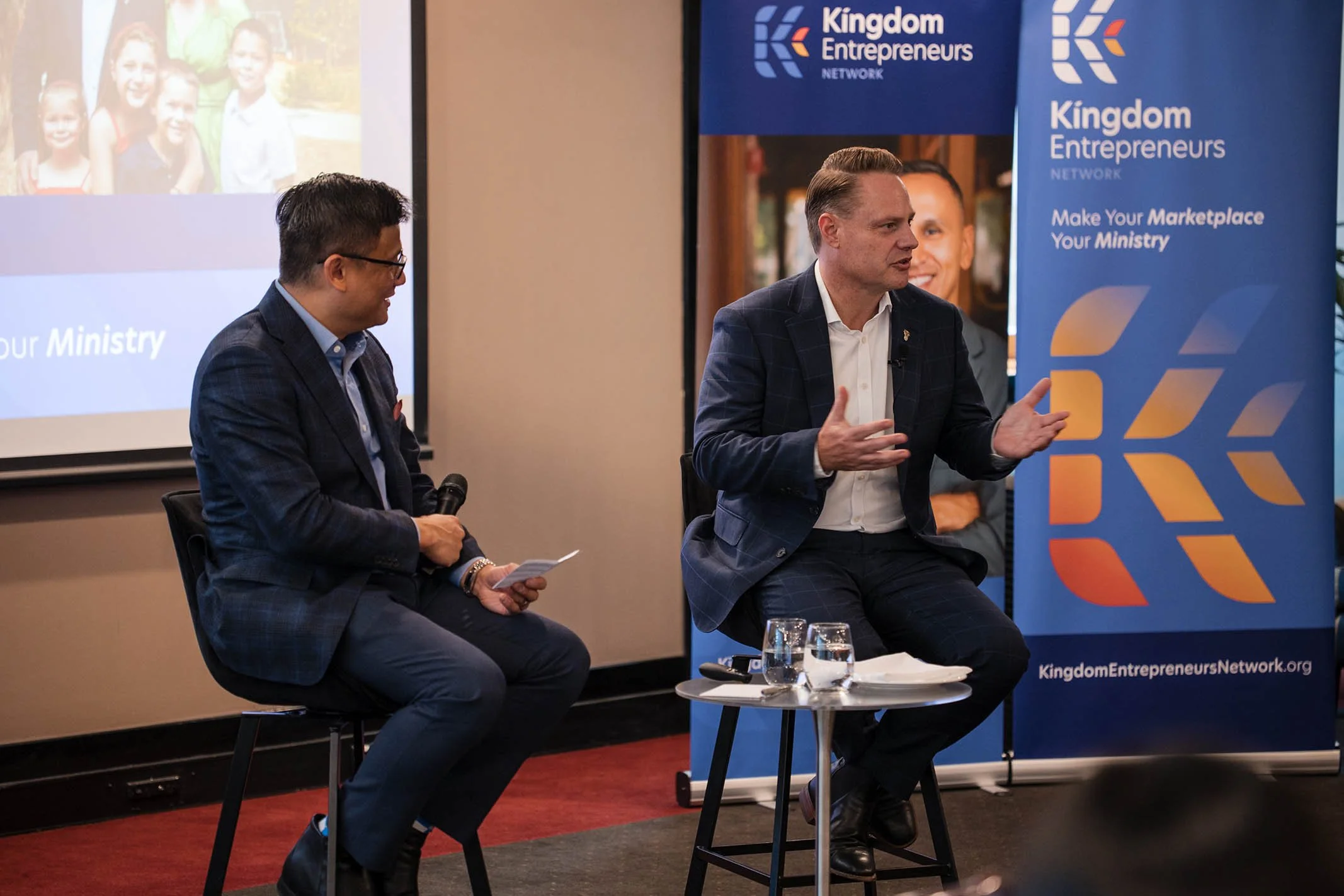 Two men seated and speaking at a Kingdom Entrepreneurs event, with banners behind them displaying the organization's logo and slogan. One man is holding a microphone, and there is a small table with water glasses in front of them.