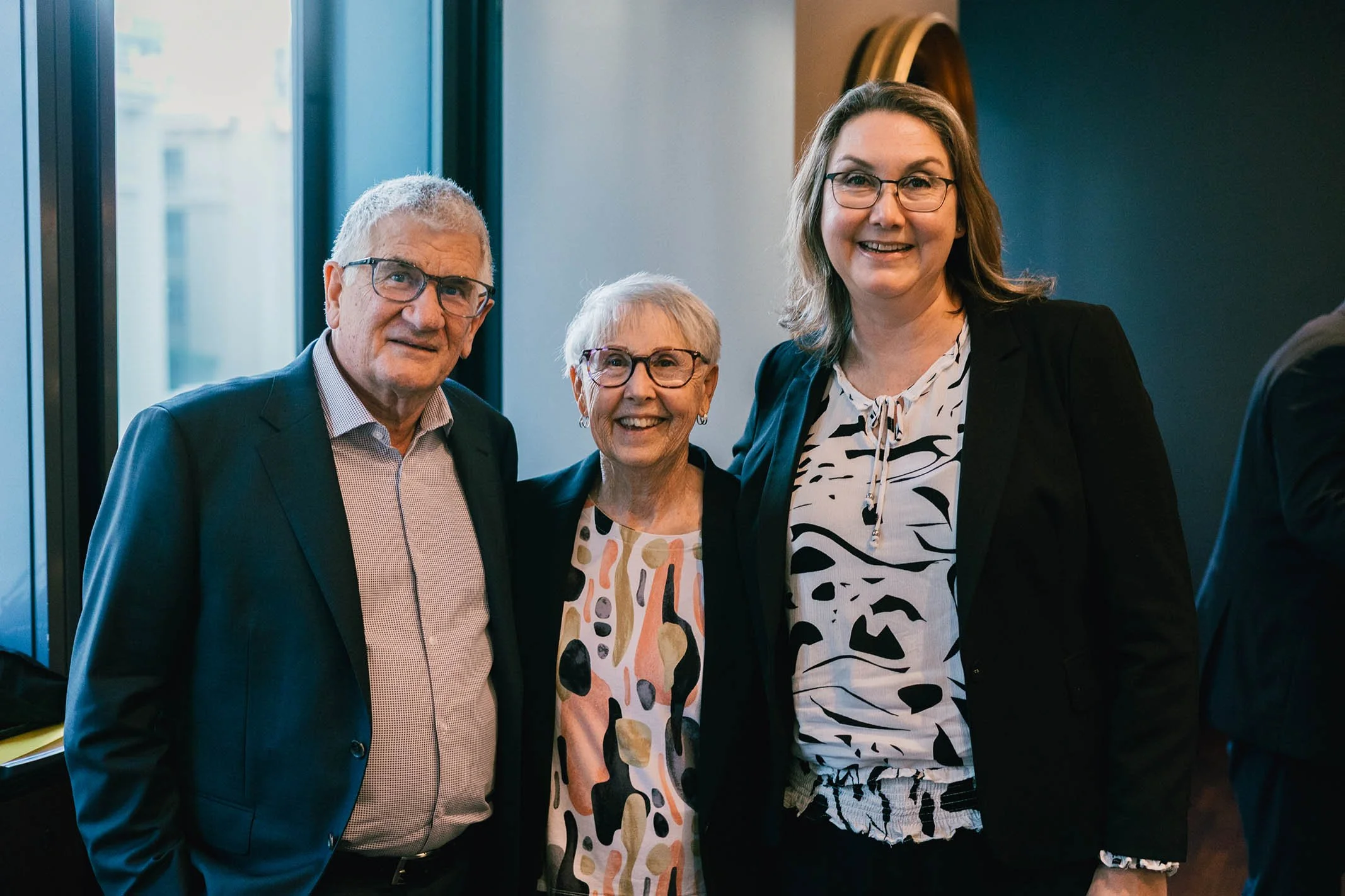 Three smiling adults, two women and one man, standing close together indoors at a social or professional event.