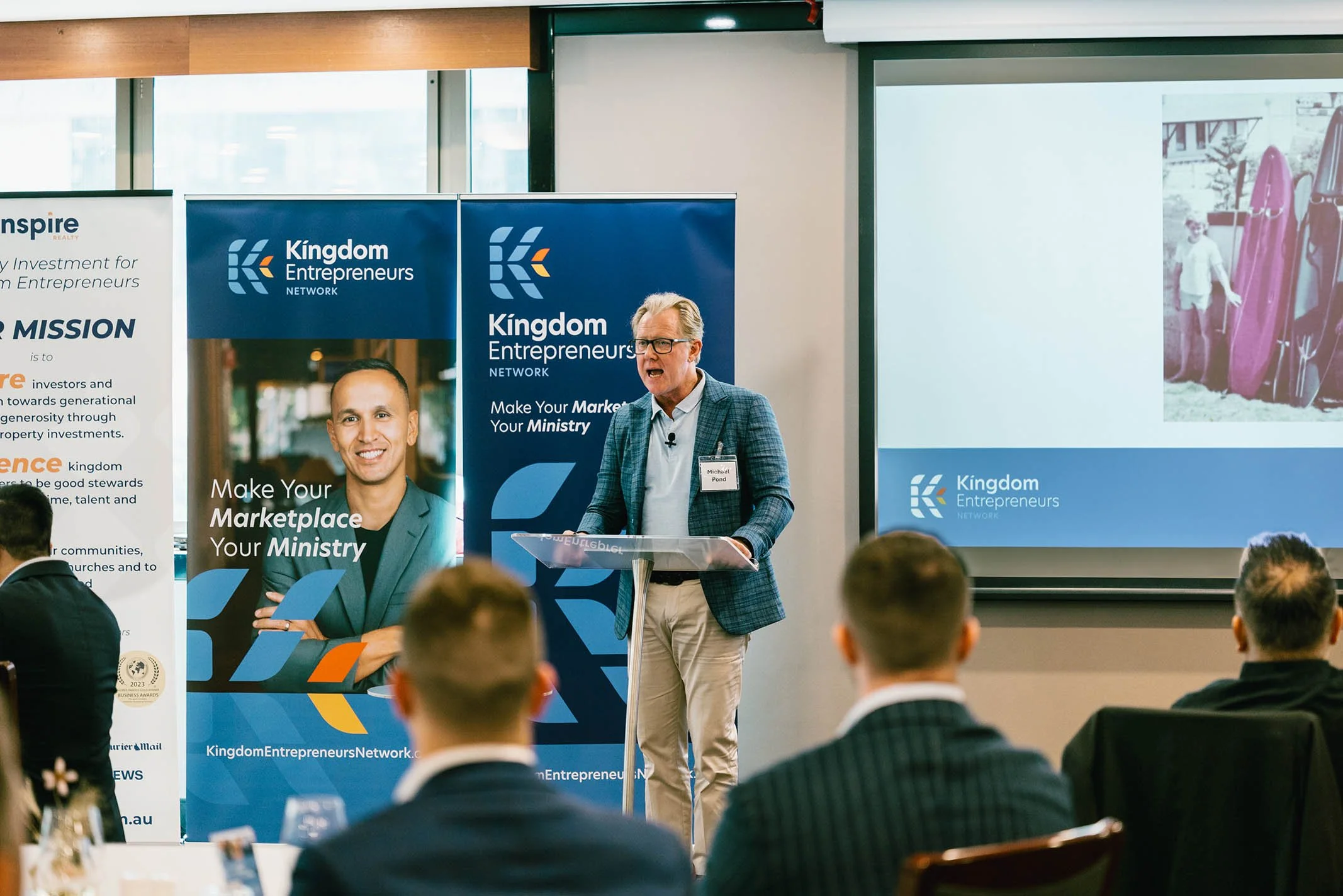 A man is giving a presentation on stage at a business conference, standing behind a lectern, with a large screen and banners behind him that display the logo and branding of 'Kingdom Entrepreneurs Network.' The audience members are seated and listeni