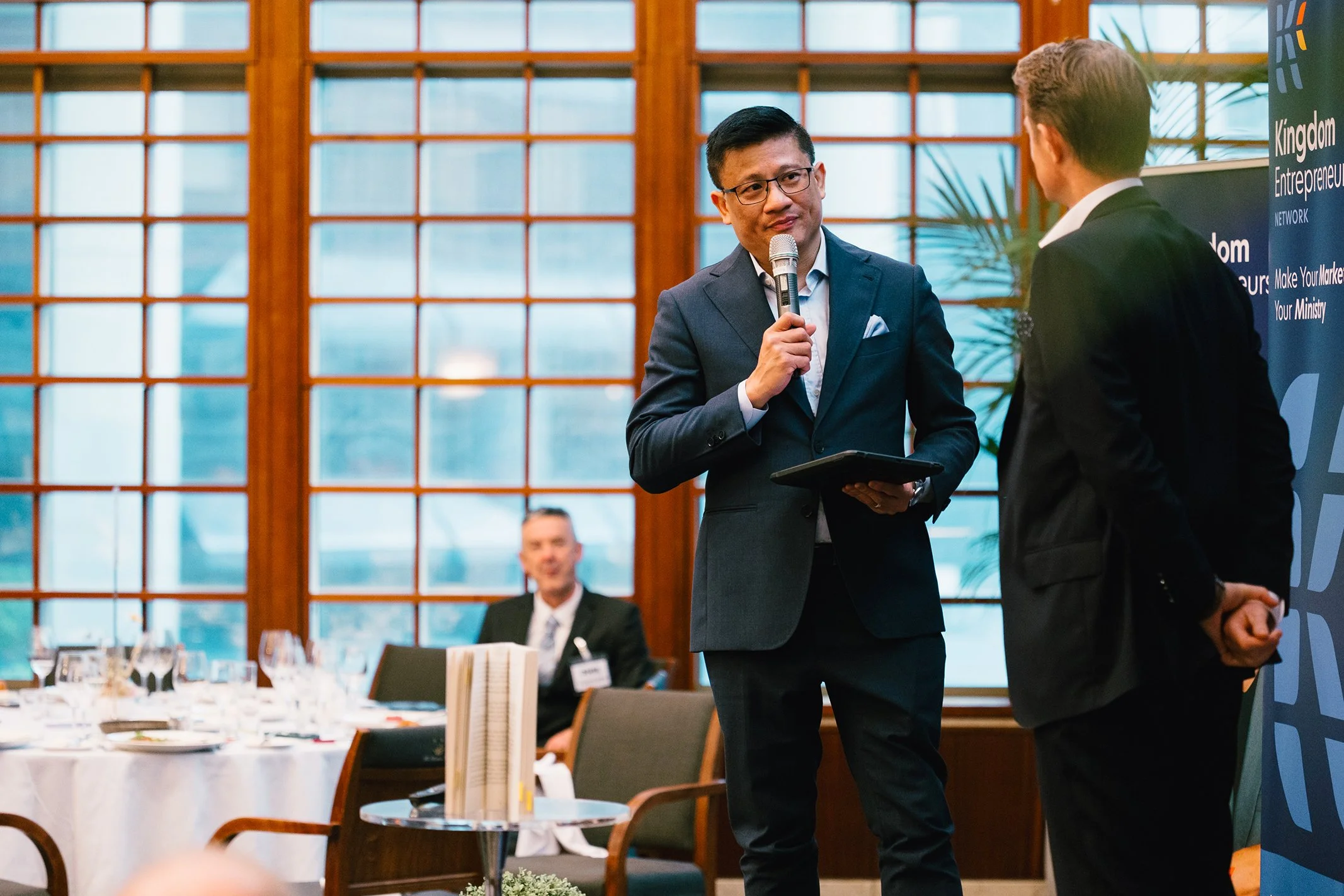 A man in a dark suit speaking into a microphone while holding a tablet, during a formal event or conference, with another man in a dark suit and a seated man in the background.