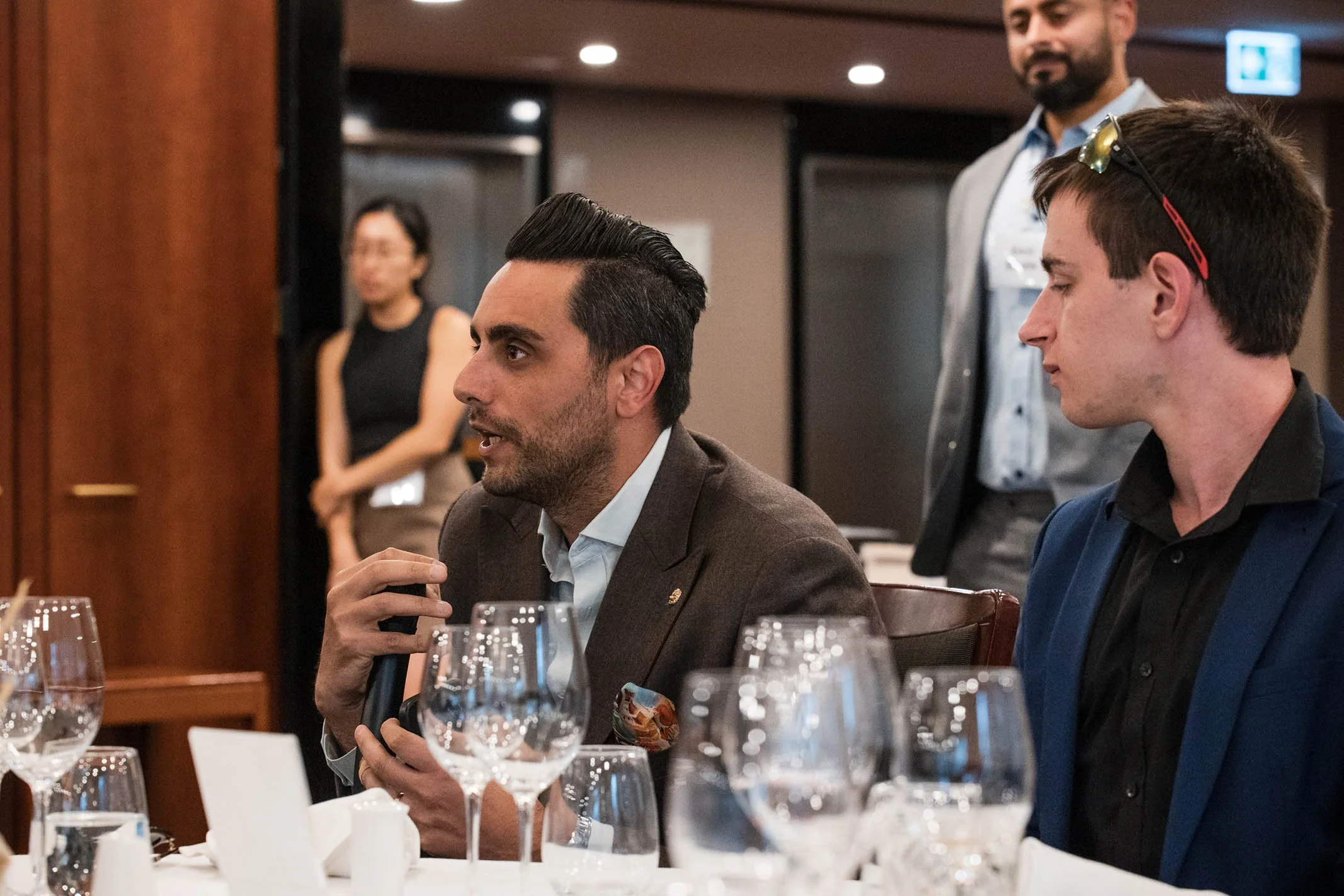 Two men sitting at a formal event, one holding a microphone and talking, with wine glasses on the table in front of them. A woman is standing in the background.