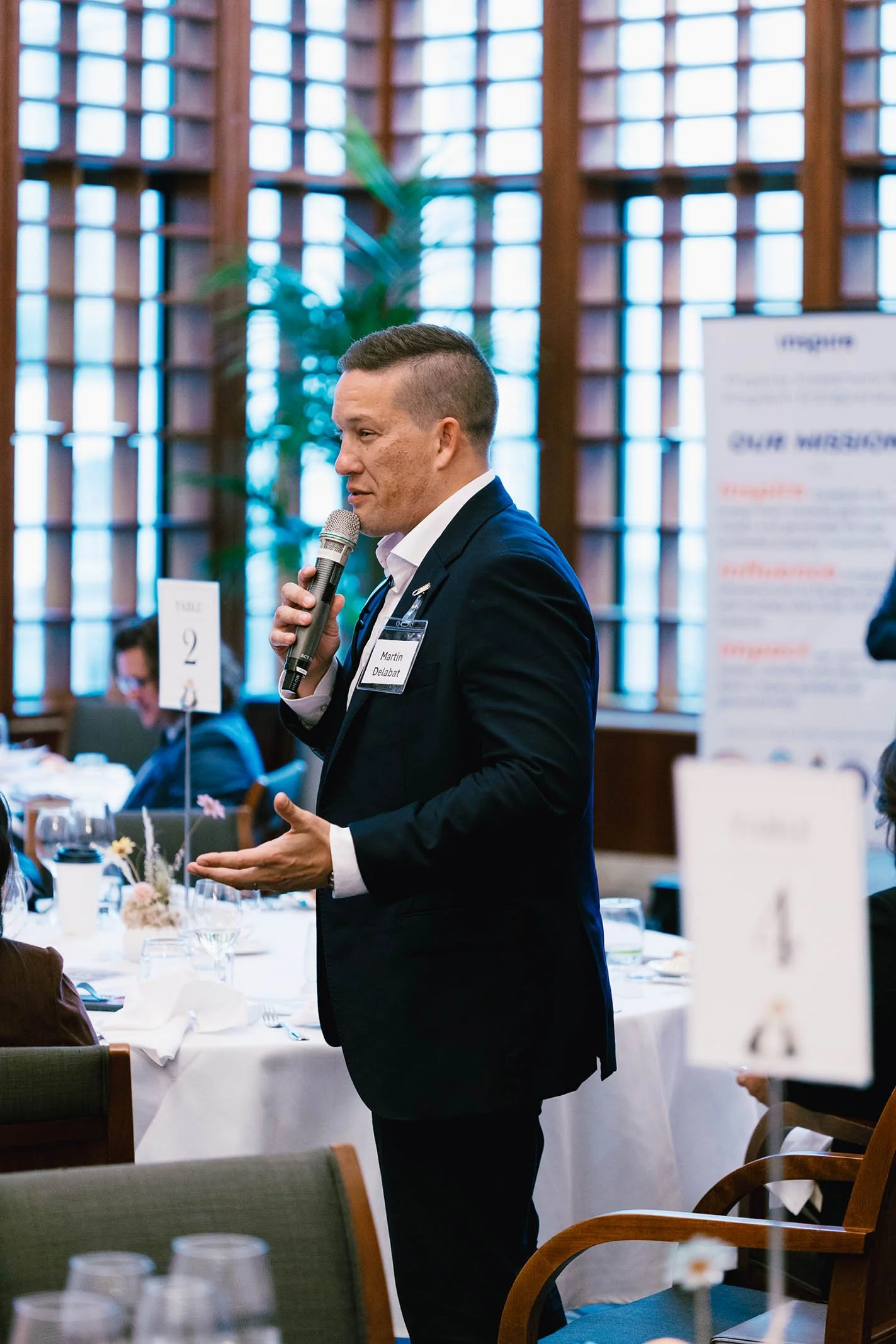 A man in a black suit with a name badge speaking into a microphone at a formal event or conference, with tables set for dining and other attendees in the background.