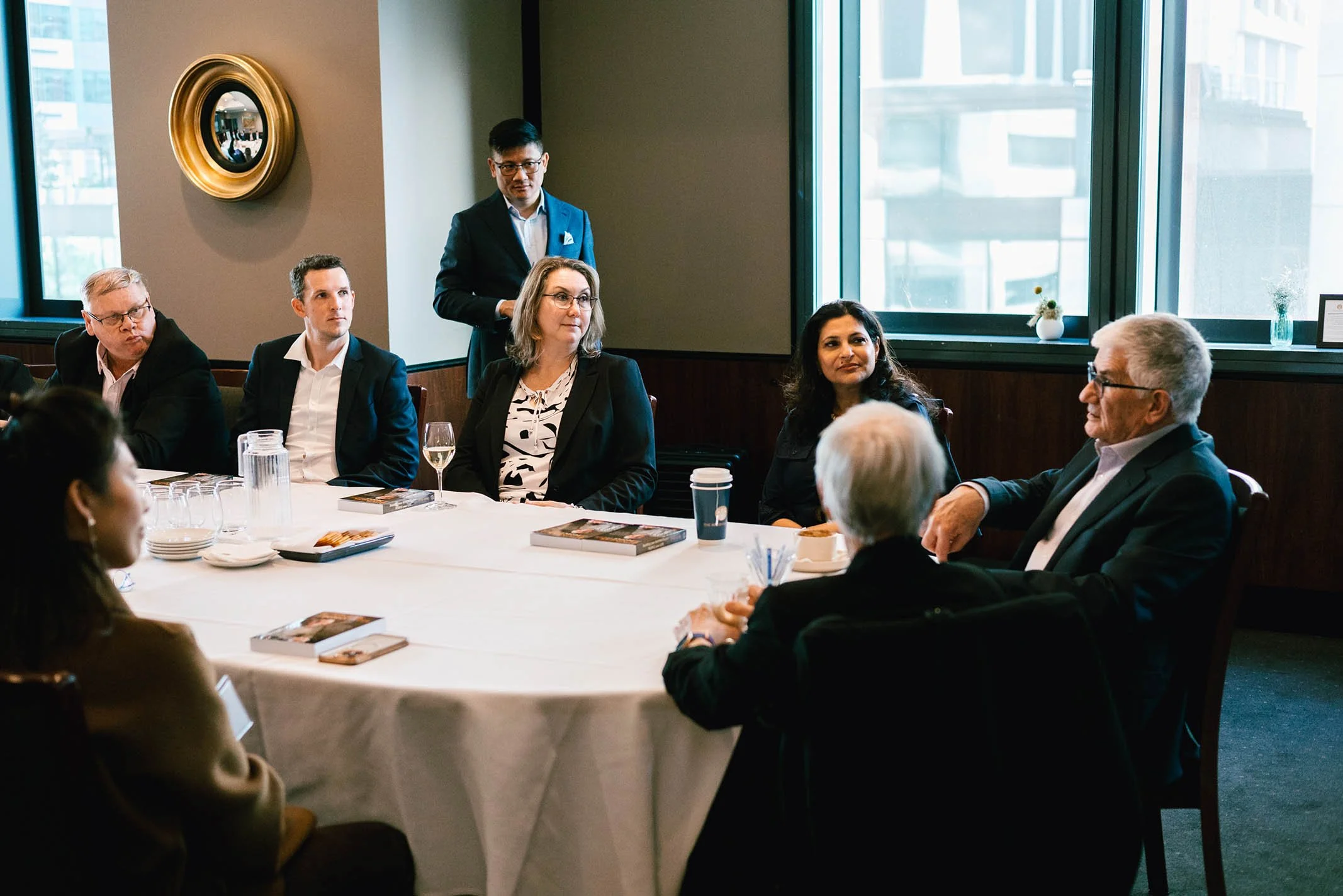Business meeting with several people sitting around a table in a conference room, one person standing, and city buildings visible through large windows.