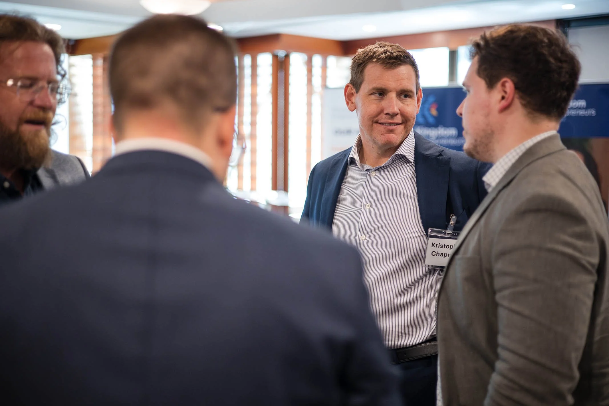 Four men in business attire having a discussion at a conference or networking event in a well-lit room with windows and banners in the background.