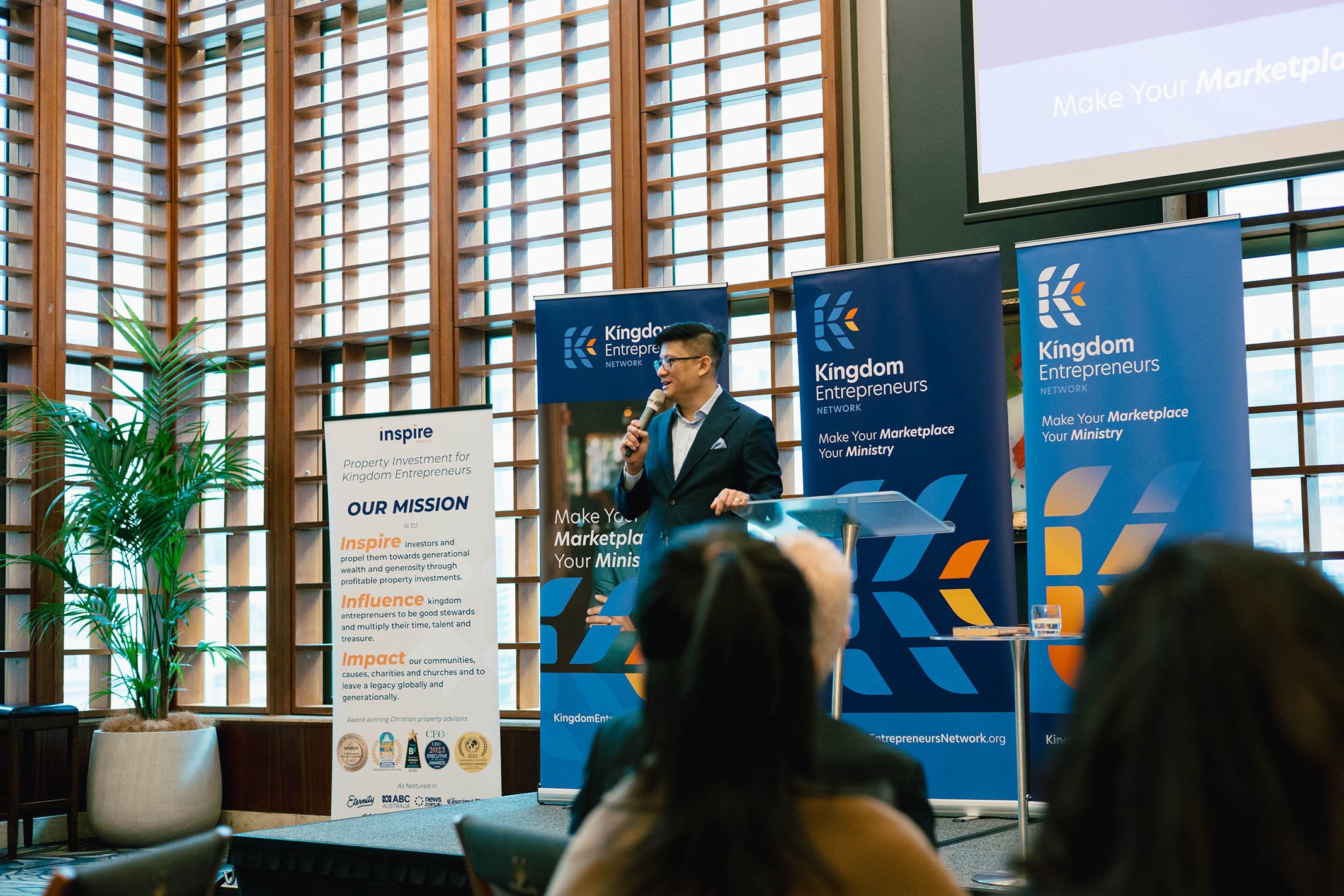 A man speaking at a podium during a conference or seminar, with banners in the background displaying 'Kingdom Entrepreneurs Network' and a white stand with the organization's mission statement.