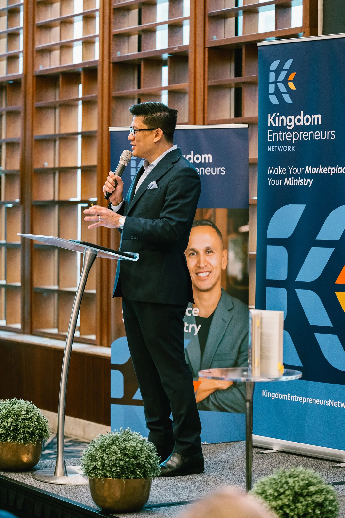 A man in a black suit and glasses is standing at a podium, speaking into a microphone at a business event. Behind him are two banners for 'Kingdom Entrepreneurs Network,' one with a large photo of a smiling man in a gray suit. The setting appears to 