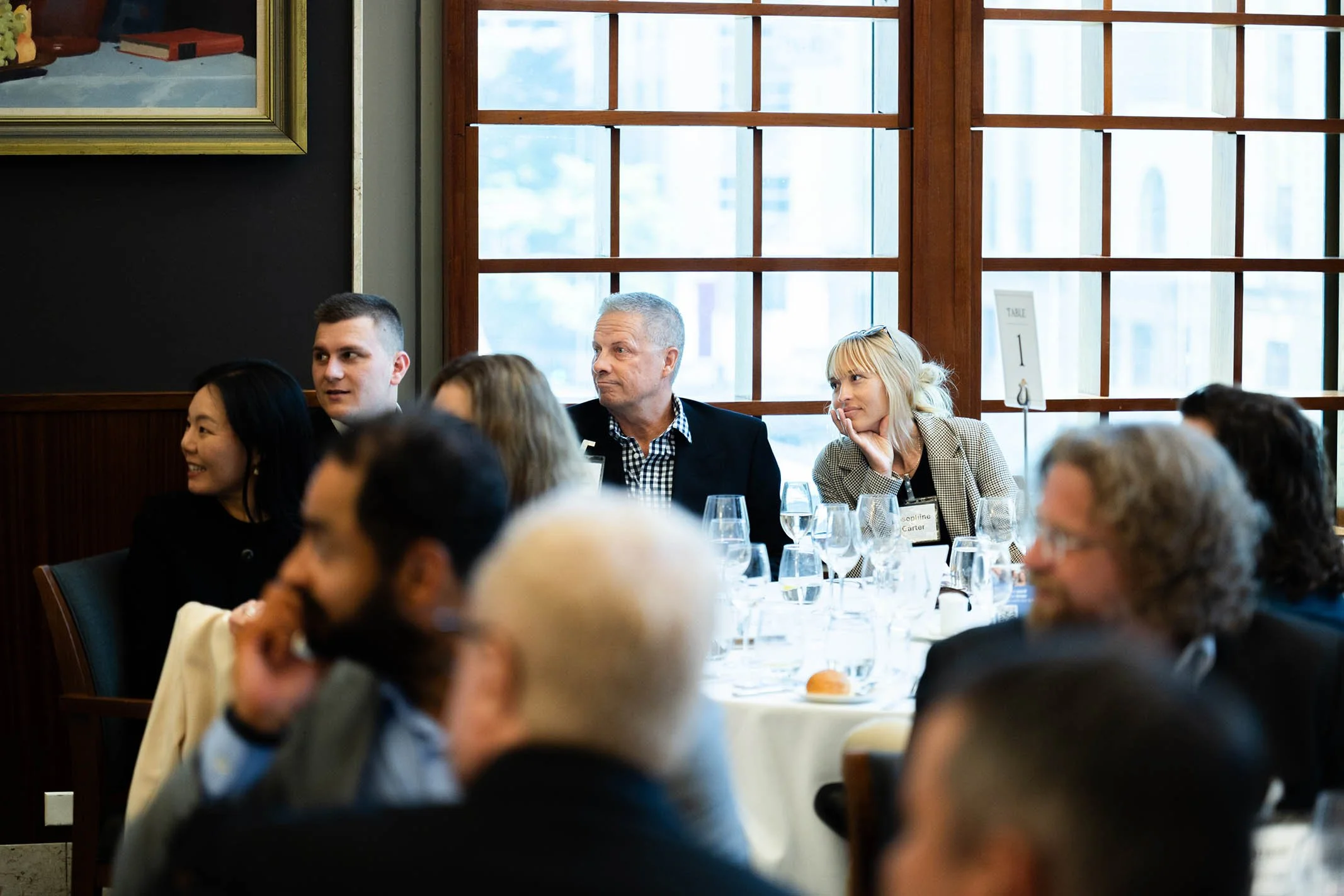 People seated at a round table during a formal event or meeting in a restaurant or conference room, with large windows in the background.