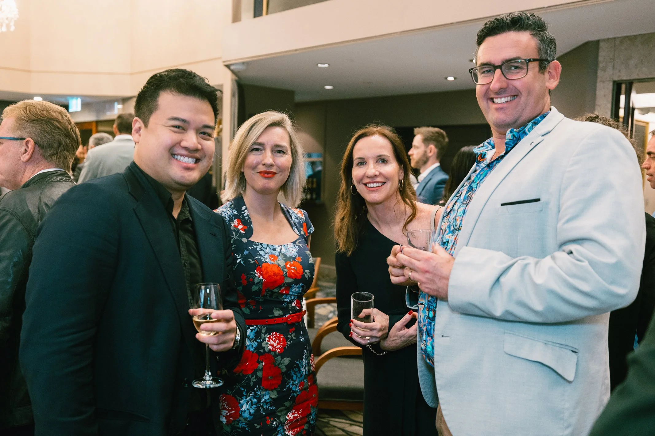 Four people at a social event, smiling, holding drinks, standing in an indoor venue with other guests in the background.