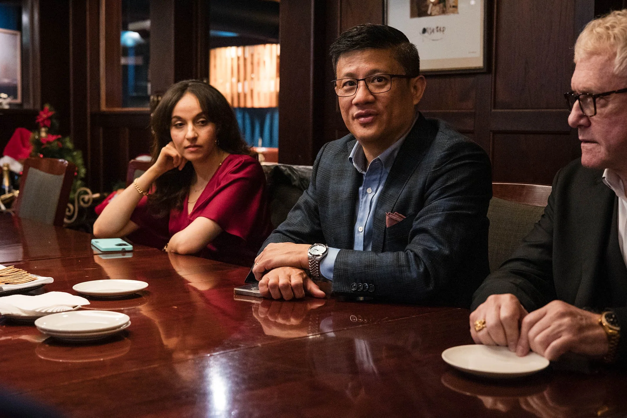 Three people sitting at a polished wooden table in a restaurant, Christmas decorations visible in the background, including a small Christmas tree with red ribbons and Santa hats, with plates and napkins on the table.