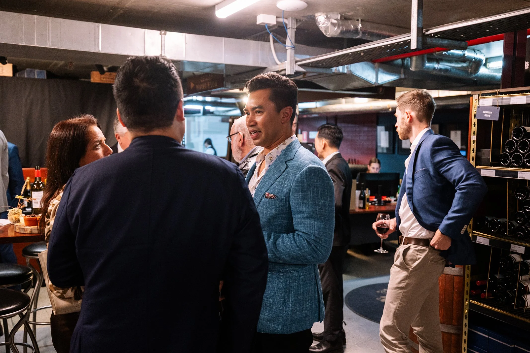 People socializing and mingling at a bar or wine tasting event, some holding glasses of red wine, in an industrial-style venue with shelves of wine bottles.