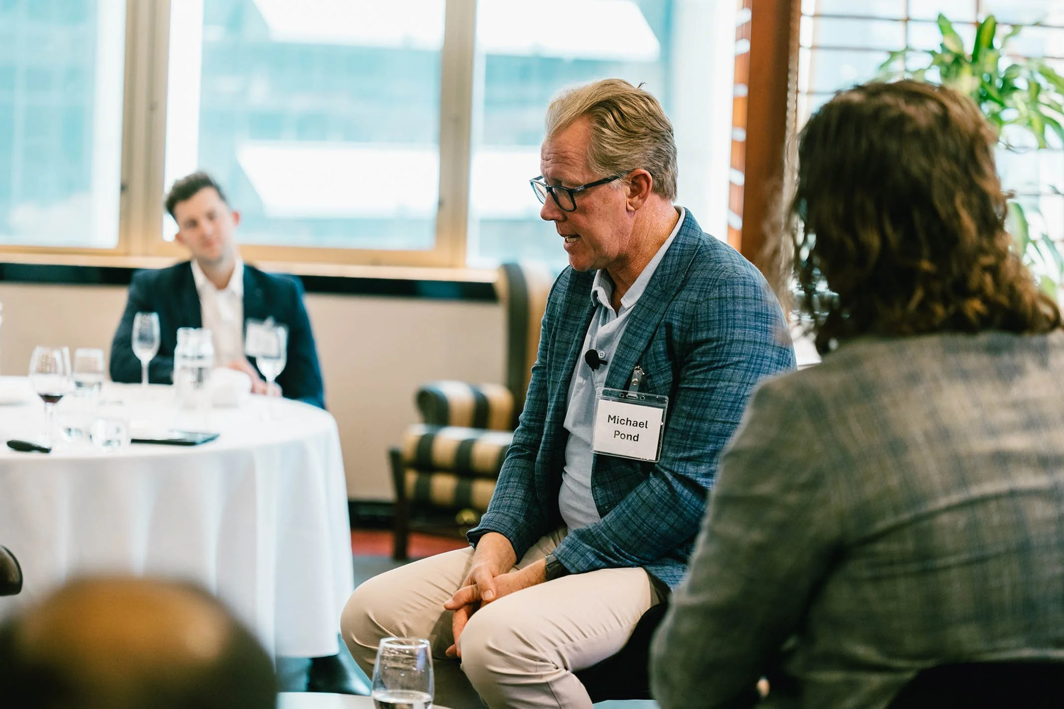 A man named Michael Pond with glasses and a name badge, wearing a blue checkered blazer, speaking at a panel or meeting, with two people visible in the background.