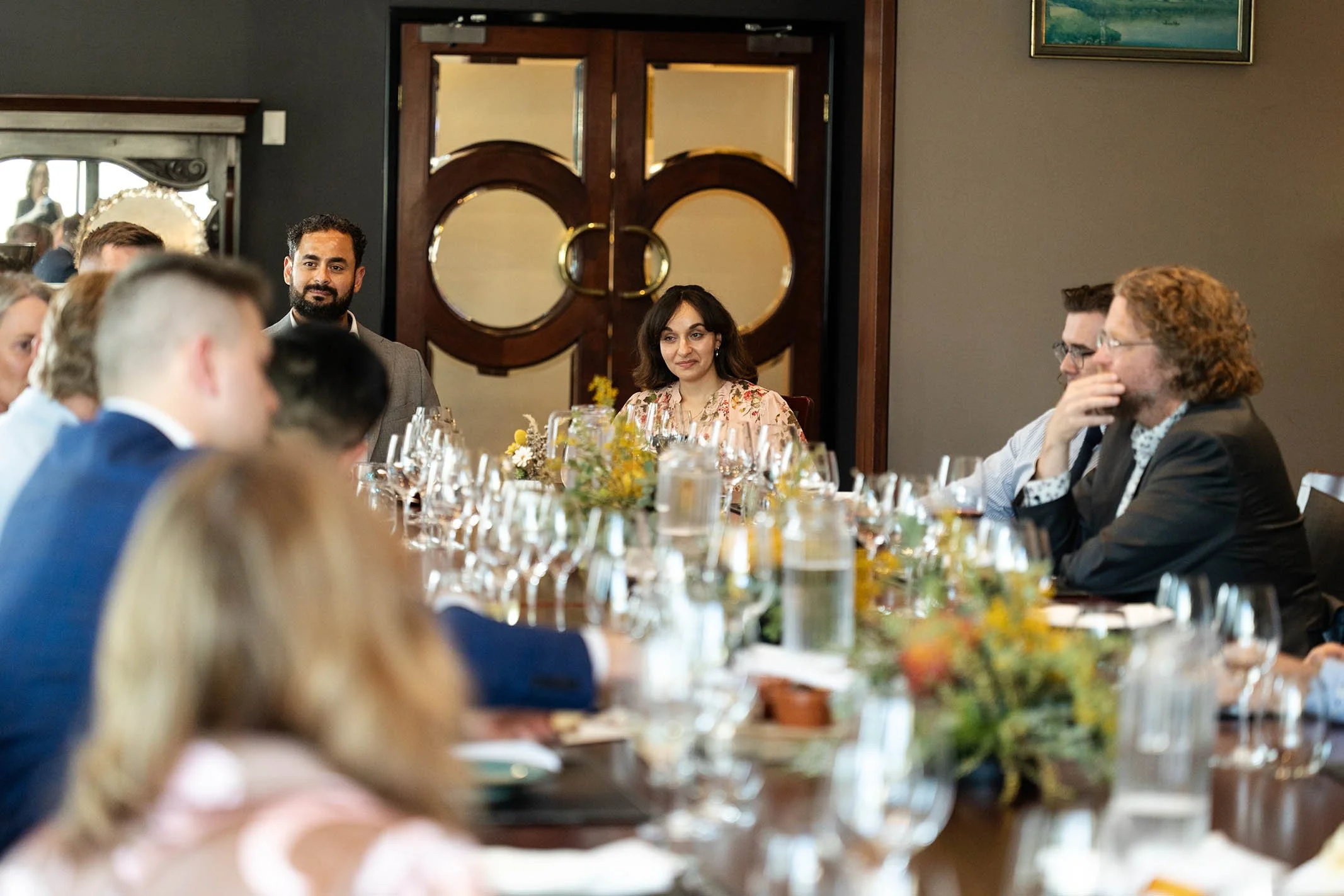 A group of people sitting around a dining table during a formal gathering or event, with some looking attentively at the center while others are engaged in conversation, decorated with flowers and glassware, in a room with dark walls and decorative w