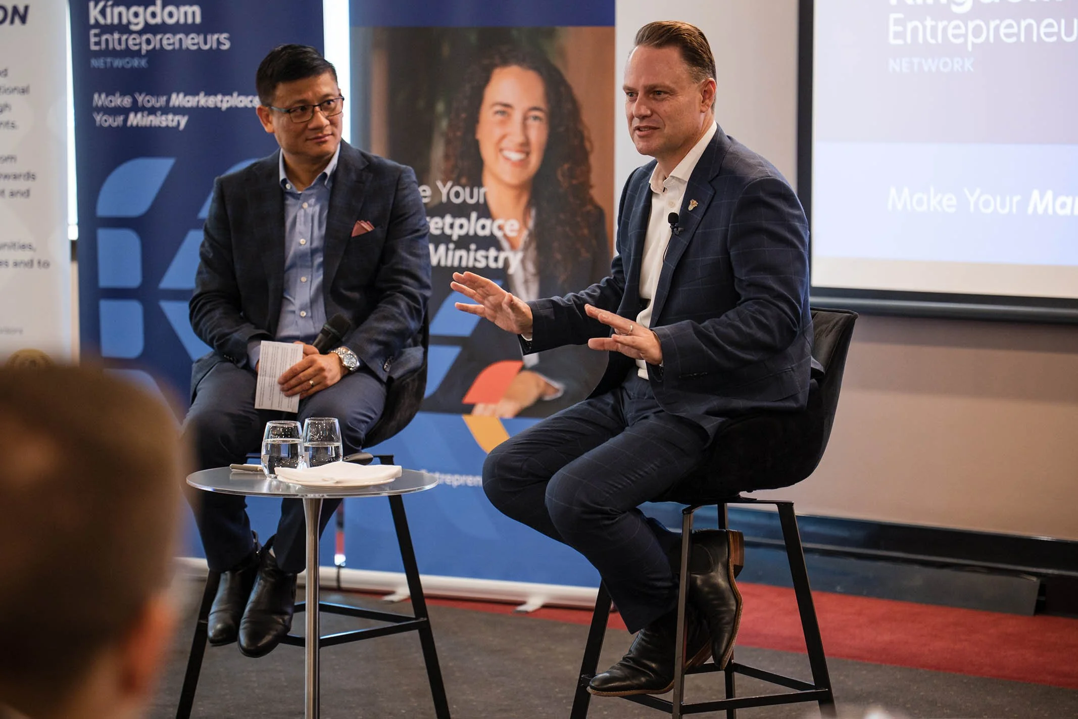 Two men in suits participating in a panel discussion or interview at an event, sitting on high chairs with a small table with water glasses between them, a microphone on one man, and a projected presentation background with banners and images behind 
