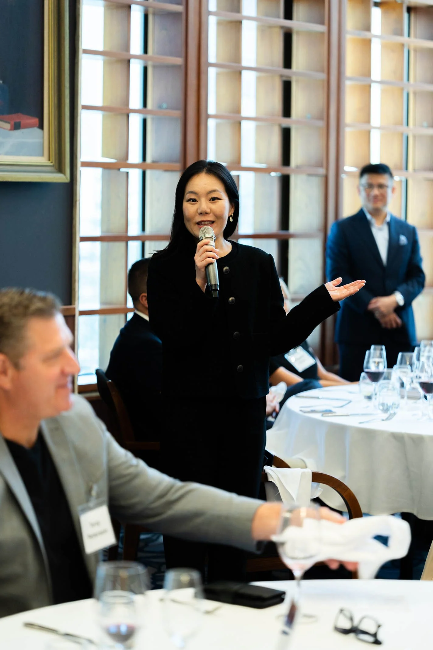 Woman speaking into a microphone at a formal meeting or event, with seated attendees and a man standing in the background.