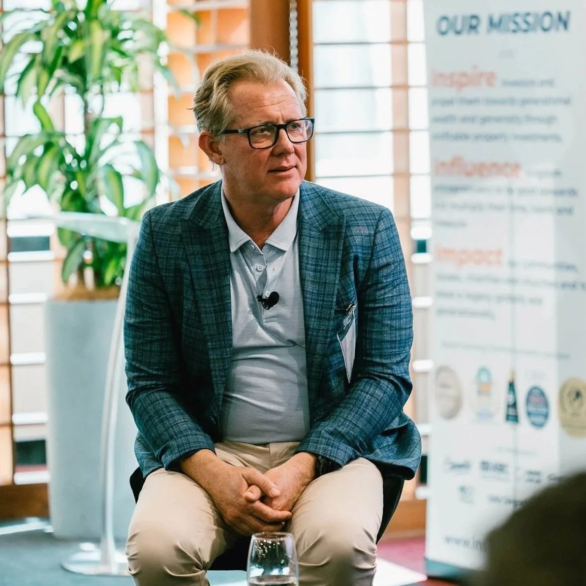 An older man with gray hair, glasses, wearing a blue plaid blazer and a light blue shirt, sitting on a chair during a discussion or presentation, with a blurred banner and a plant in the background.