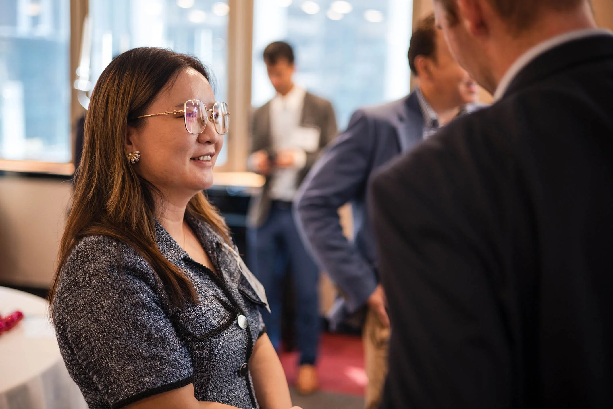 A woman with brown hair, wearing glasses and a patterned blouse, is smiling and talking to a man in a black suit at a professional event, with other people in the background.