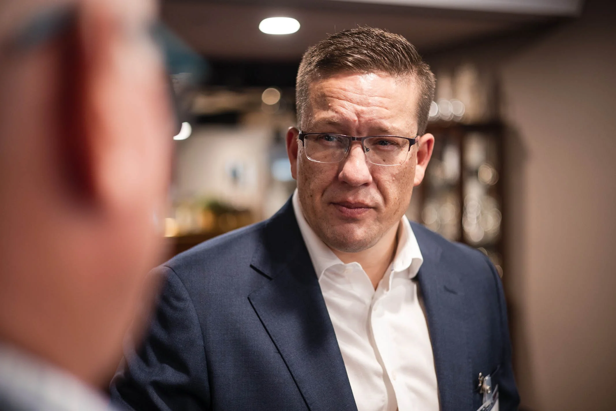 A man wearing glasses and a suit looks at another person, who is partially visible in the foreground, in what appears to be an indoor setting.
