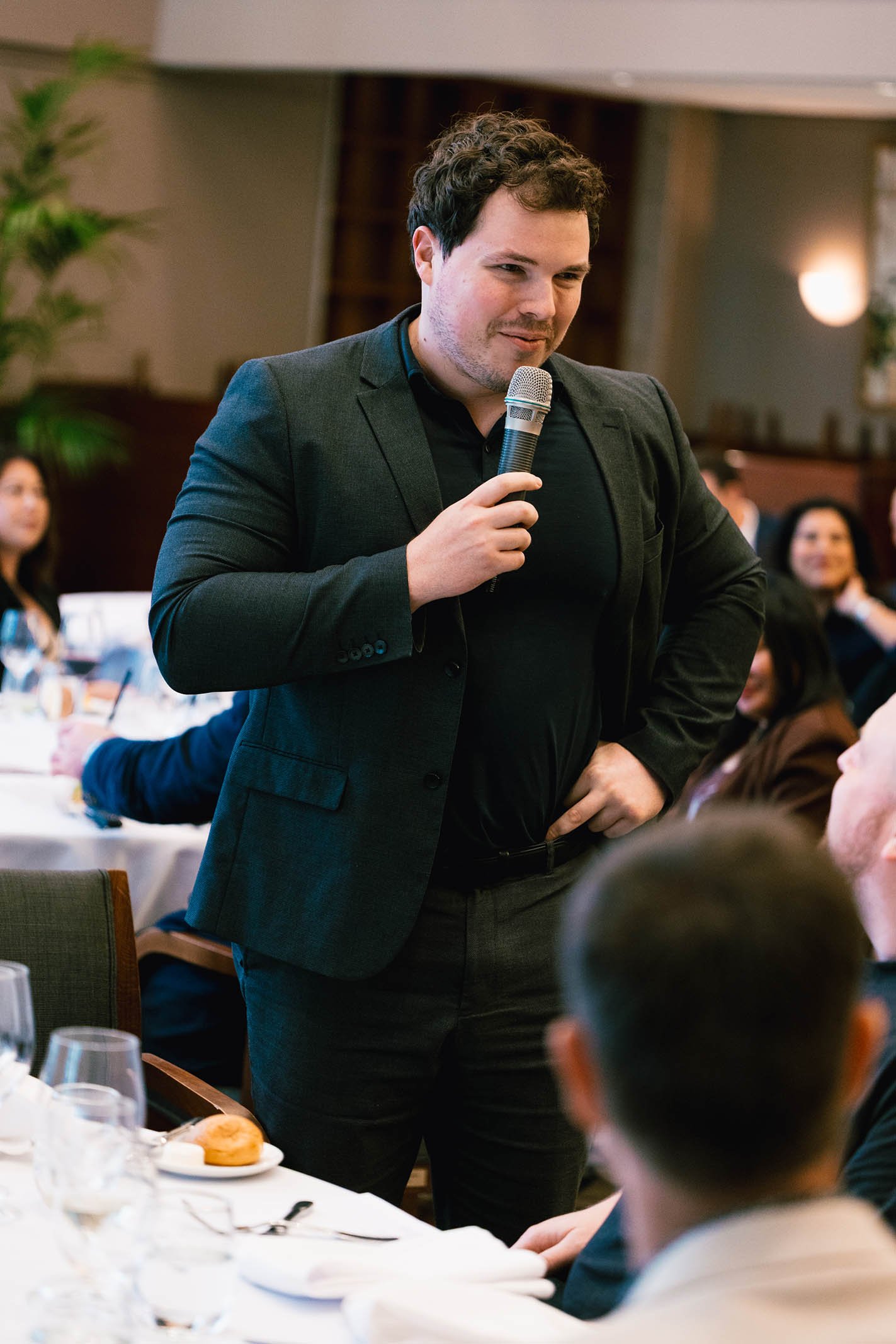 A man in a dark blazer and black shirt standing at a table, speaking into a microphone during an event. Other people seated at the table are listening, with some smiling, while the table is set with glassware, plates, silverware, and a bread roll.