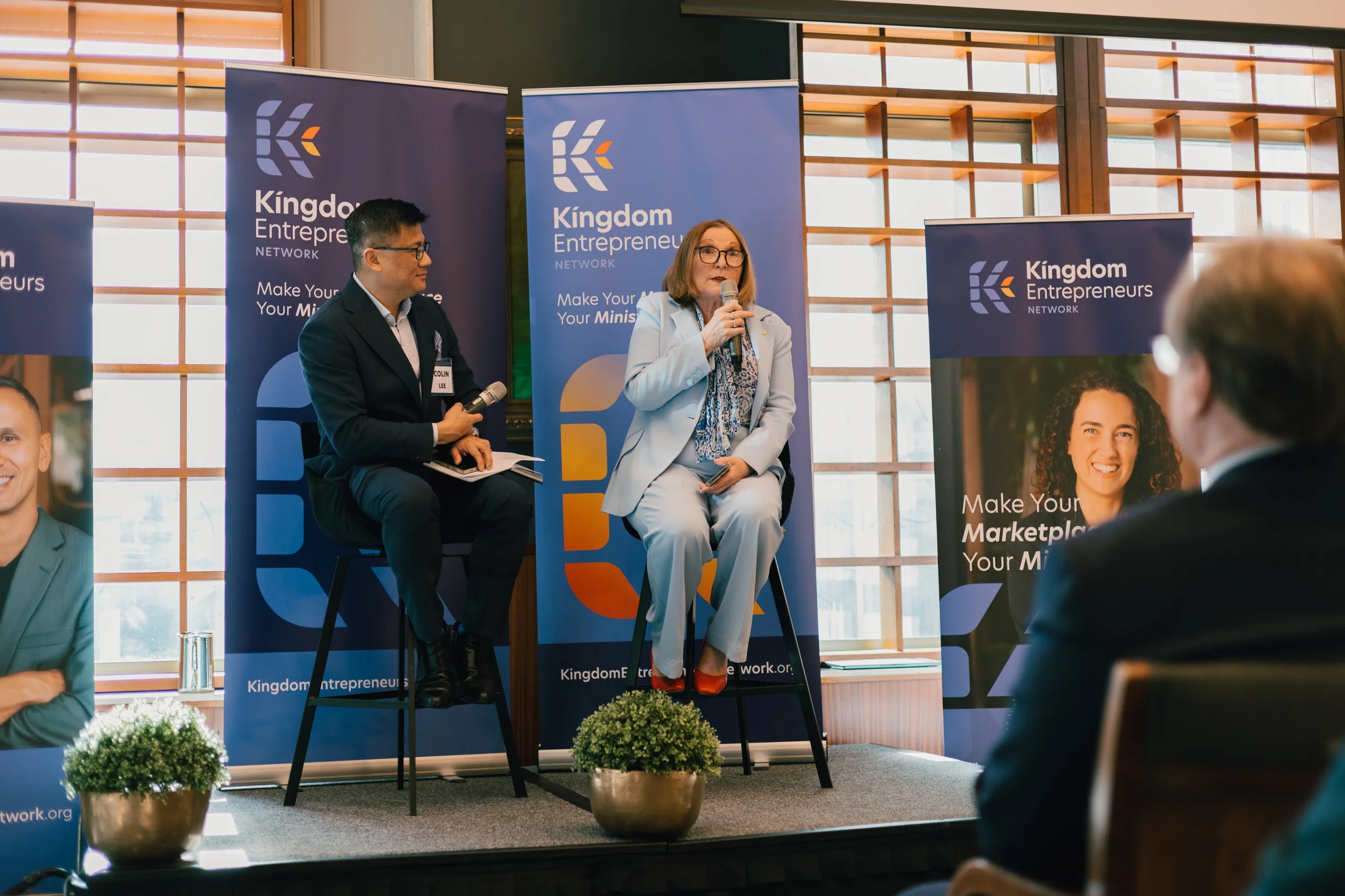 Two speakers, a man and a woman, on stage at a Kingdom Entrepreneurs Network event. The woman is speaking into a microphone, and there are blue banners with the event logo and slogan behind them. Audience members are visible in the foreground.
