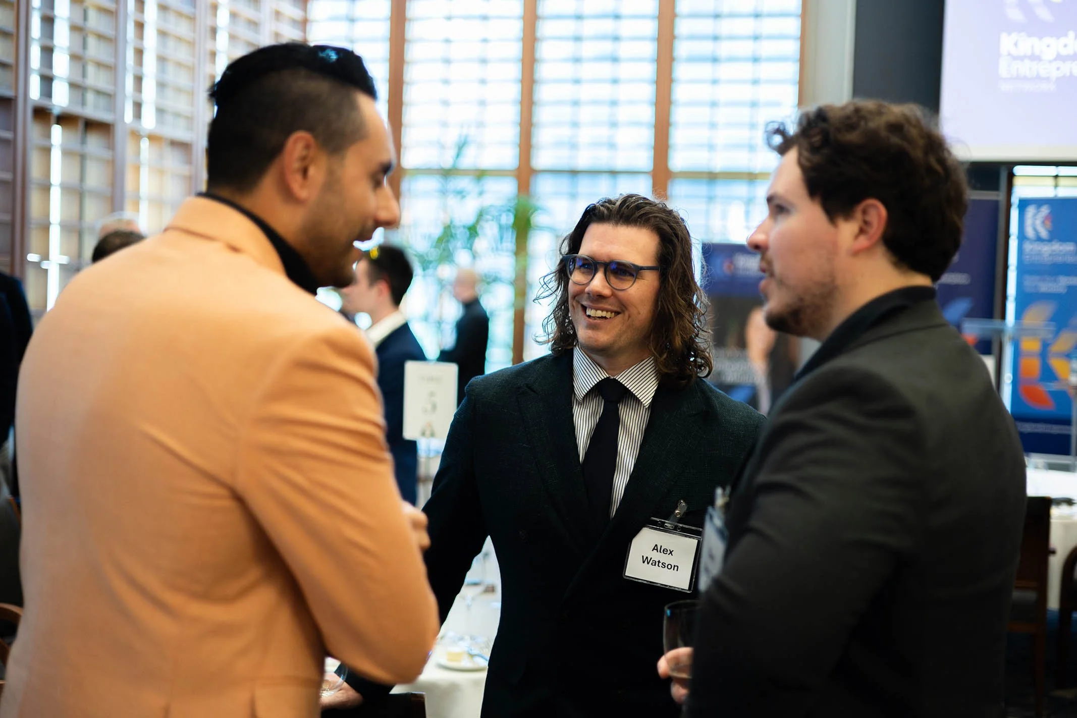 Three men in formal attire engaged in conversation at a professional networking event, with name tag visible on one of the men reading "Alex Watson."