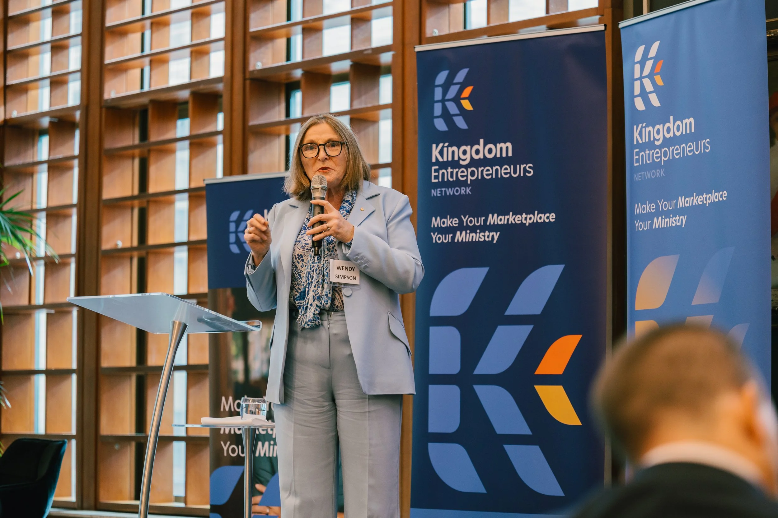 A woman with glasses and a name tag that reads Wendy Simpson is speaking into a microphone at a podium during a conference or event. Behind her are two large blue banners with the logo and text for the Kingdom Entrepreneurs Network, which includes th