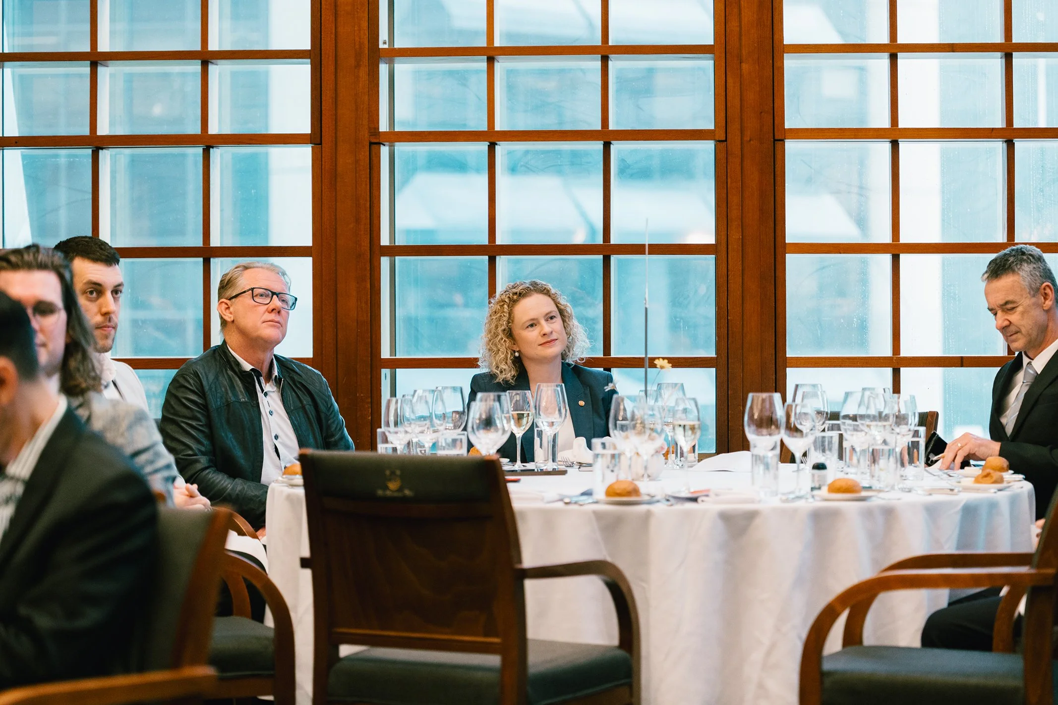 People sitting at a round table during a formal event in a room with large wooden-framed windows.