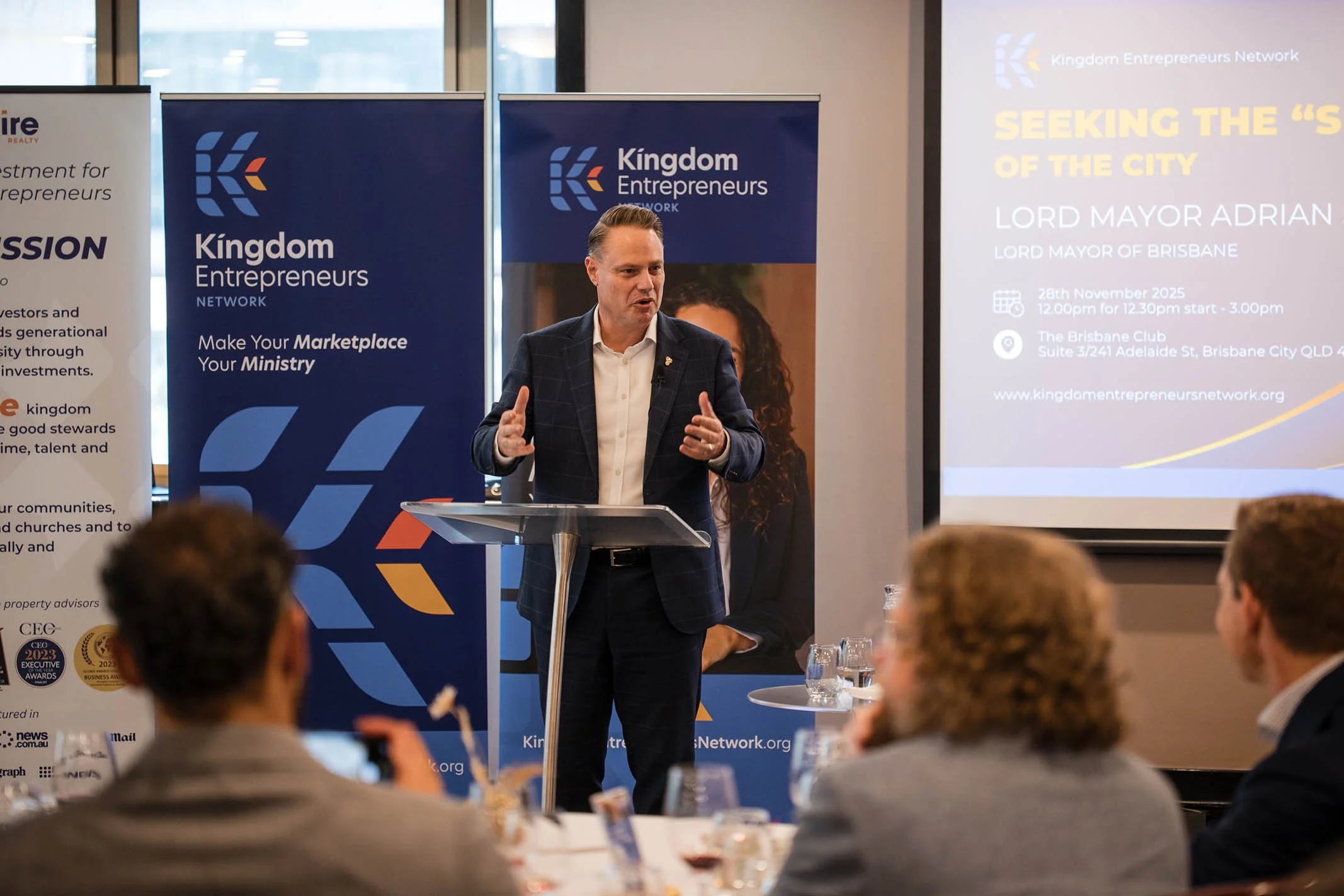 A man in a dark blazer and white shirt speaking at a conference, standing at a lectern with a microphone, with banners and a projection screen behind him in a conference room.