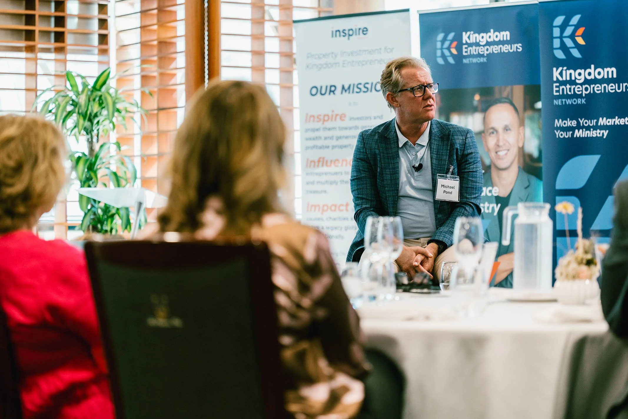 A man named Michael Pond speaking at a conference with a Kingdom Entrepreneurs banner in the background, seated at a table with others, in a bright room with wooden blinds and a plant.