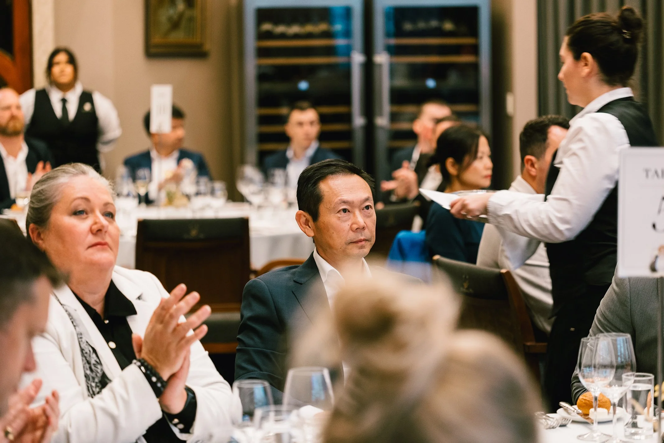 A formal dinner event with several people seated at tables, listening to a woman in a black vest and white shirt who is speaking to the group. Some guests are clapping while others are attentively listening.