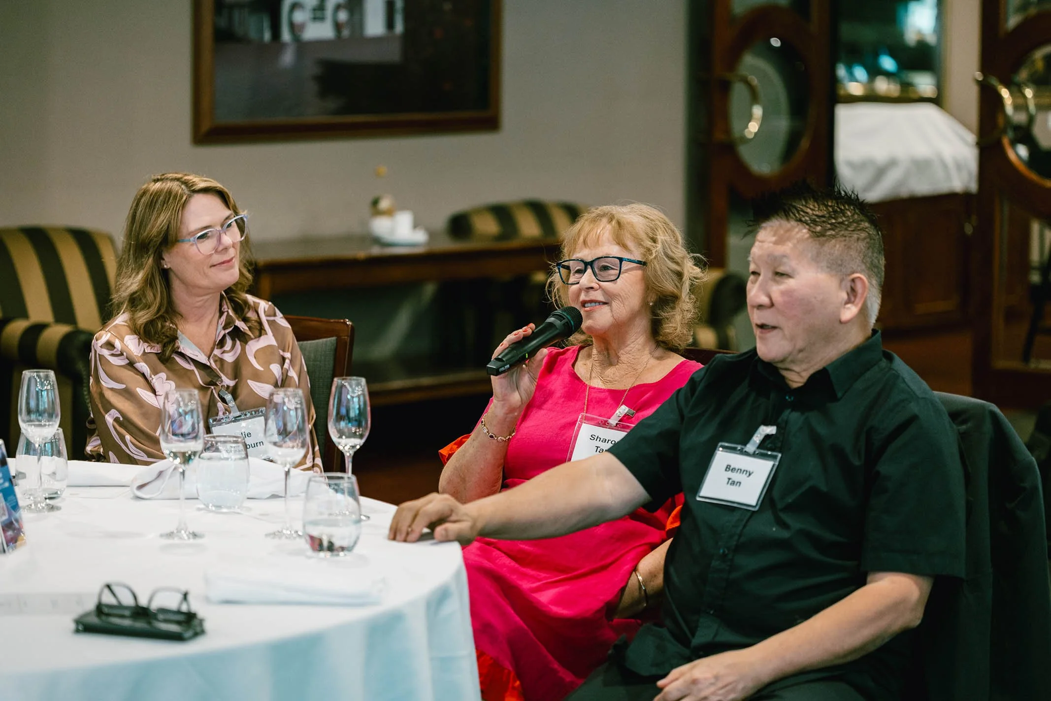 Three people seated at a round table in a restaurant or banquet hall, with one woman speaking into a microphone, while the other woman and man listen. All are wearing name tags, and the table is set with multiple glasses, napkins, and a pair of glass