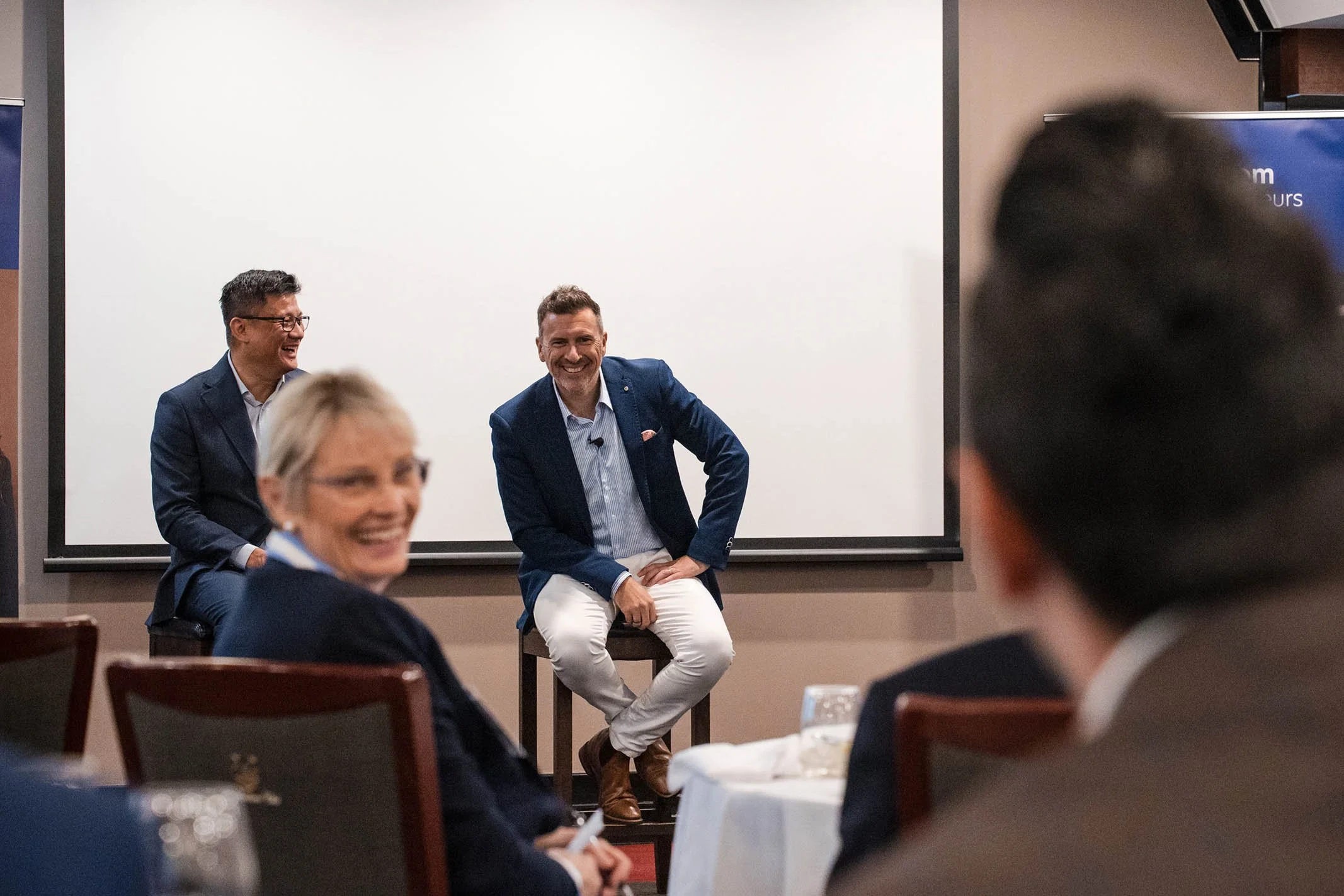 Two men are sitting on a stage and speaking to an audience at a conference. One man is smiling, wearing a dark blue blazer and white pants, and the other is laughing, wearing glasses and a navy blazer. The audience members are seated at tables, one w