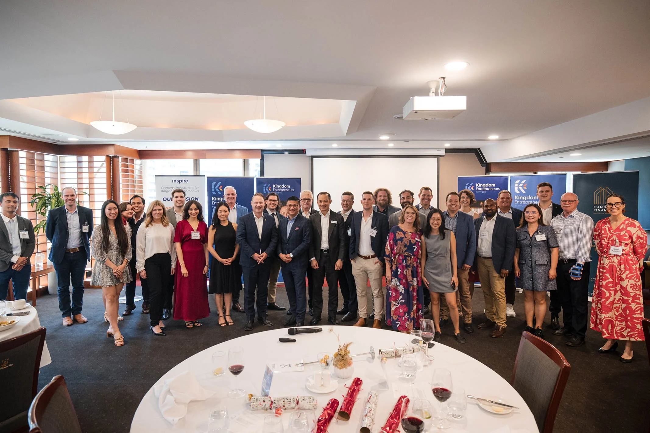 Group of professionally dressed people standing together in a conference room with banners in the background and a table with wine glasses and wrapped gifts in the foreground.