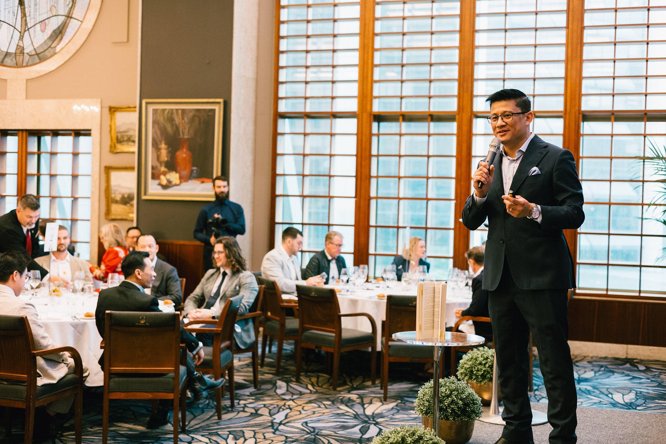 A man in a black suit is giving a speech or presentation using a microphone in a formal setting with several seated attendees in business attire, large windows, and artwork on the walls.