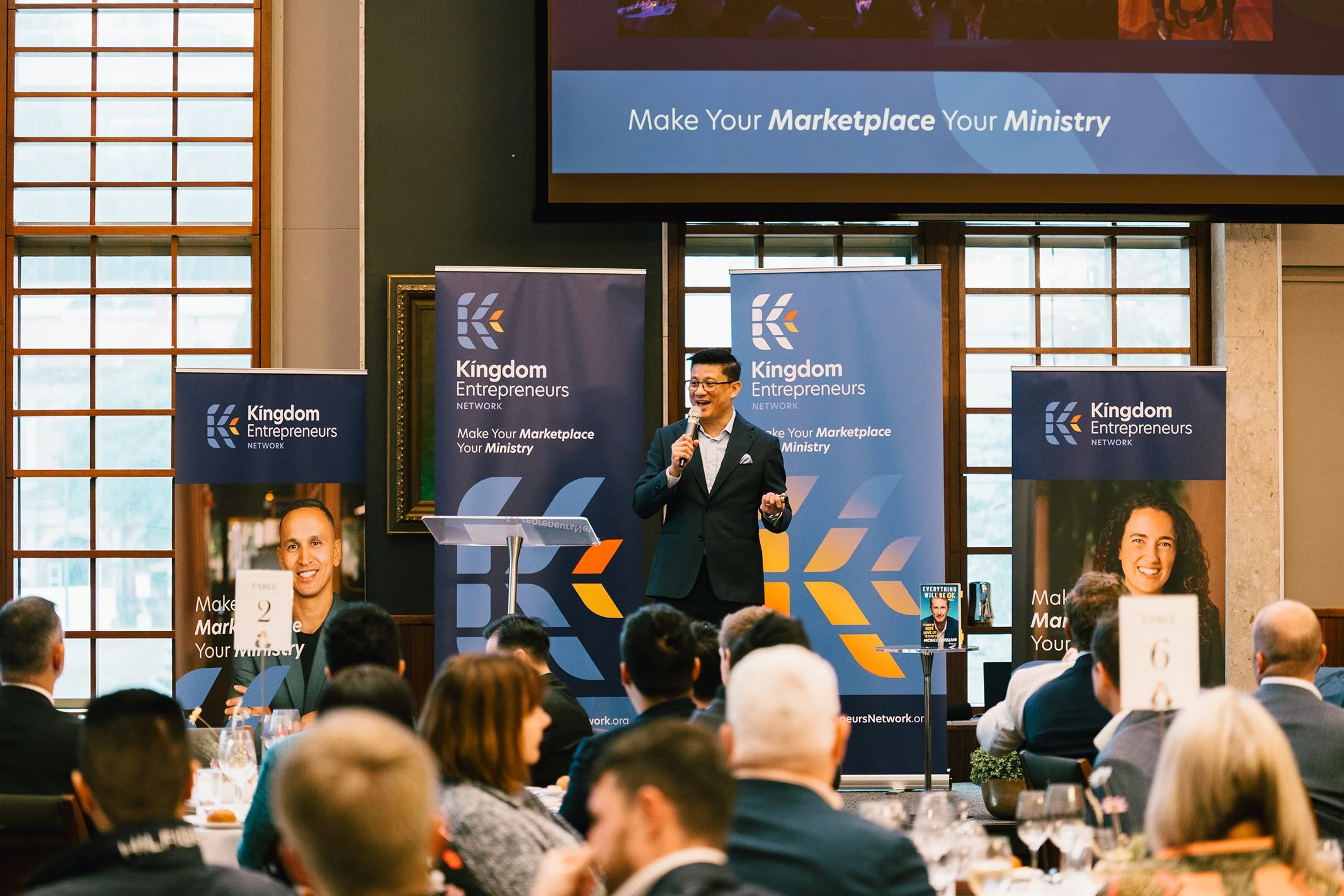 A man in a suit giving a presentation on stage at a conference, with banners displaying 'Kingdom Entrepreneurs Network' and the slogan 'Make Your Marketplace Your Ministry' behind him. The audience is seated at tables.