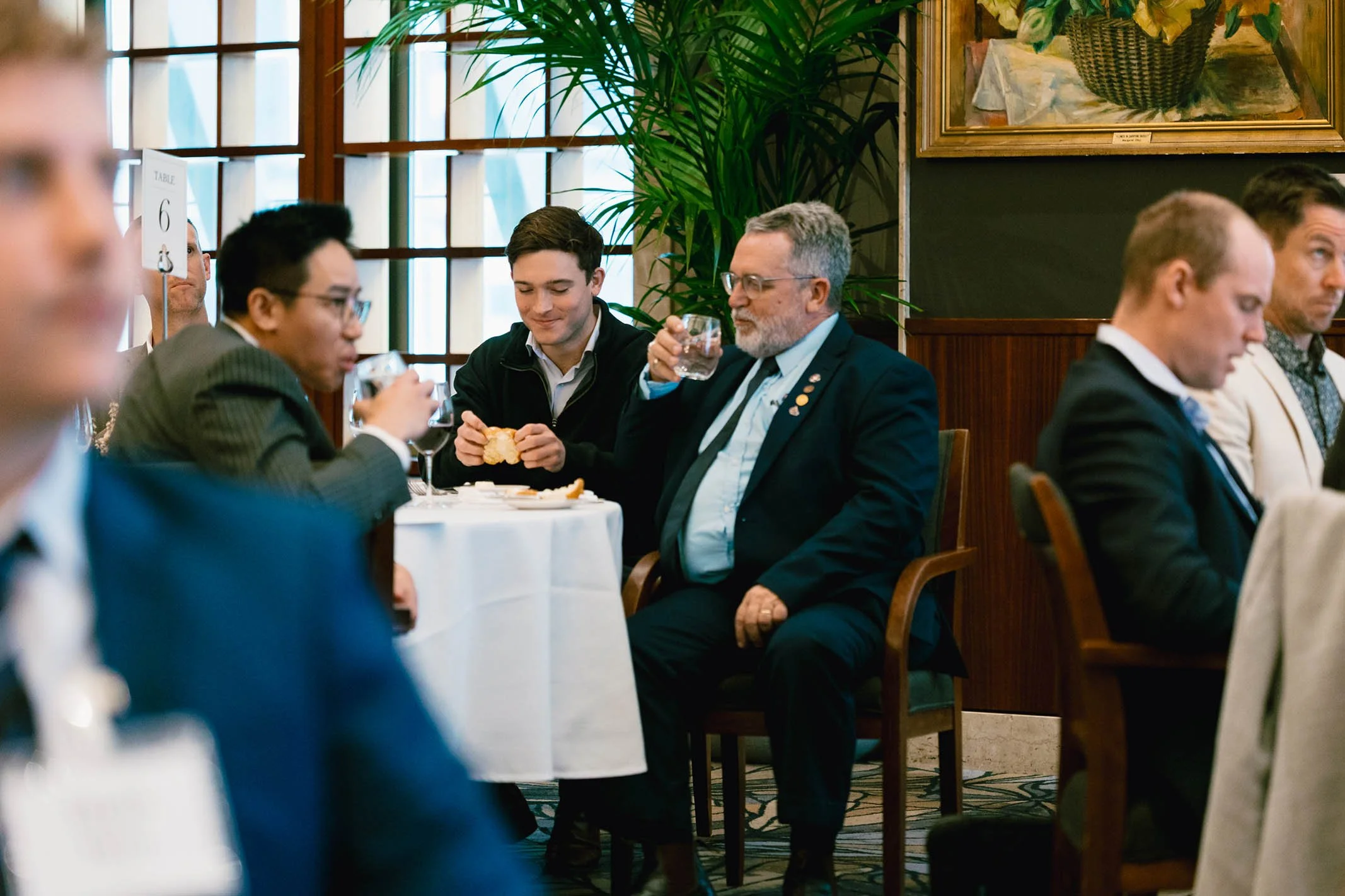People sitting at a round table in a restaurant or banquet hall, some eating and others drinking, with large windows and a plant in the background.