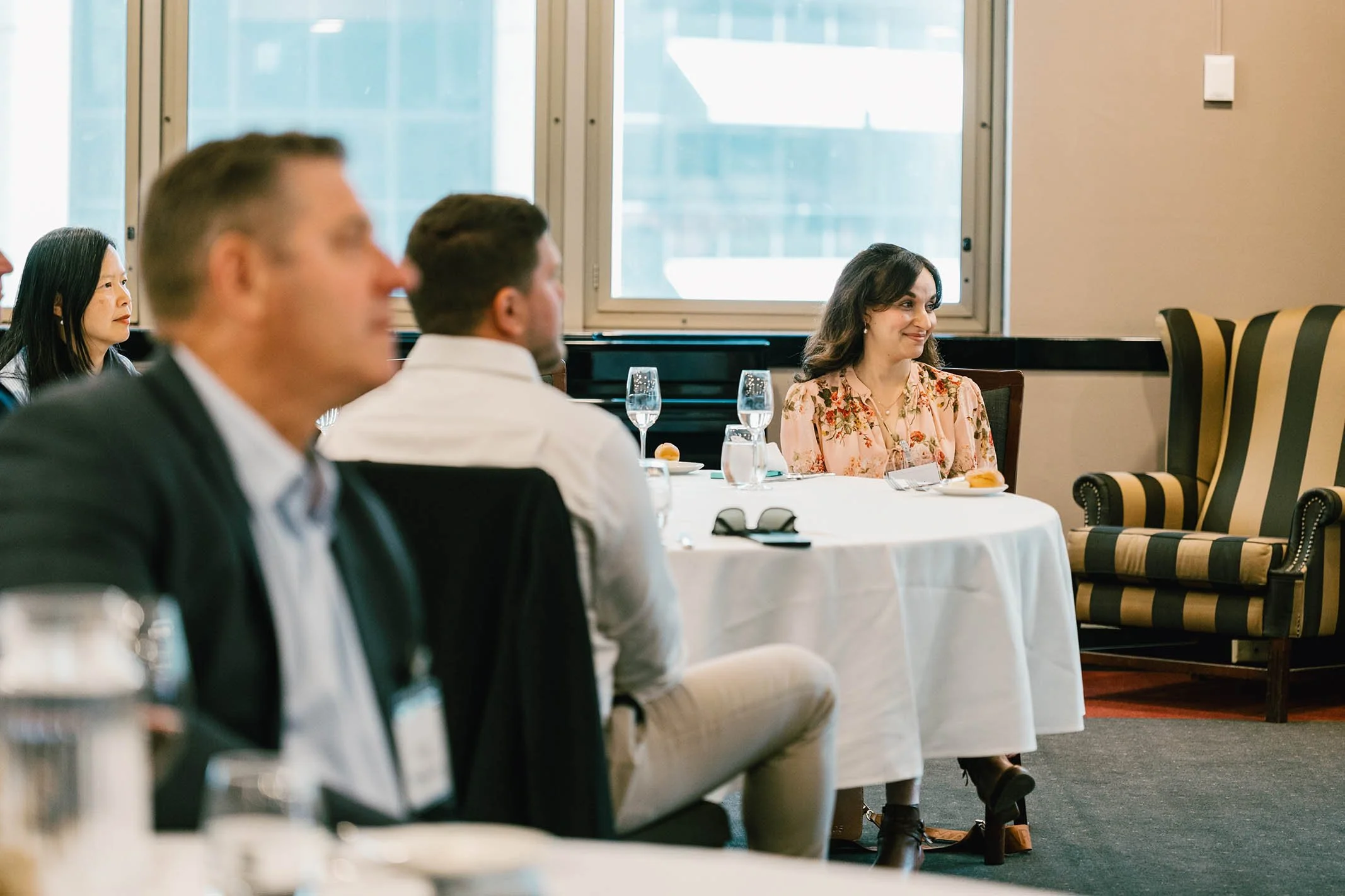 People attending a formal event or meeting seated around a table with glasses of water and plates, with a woman in floral attire smiling in the background.