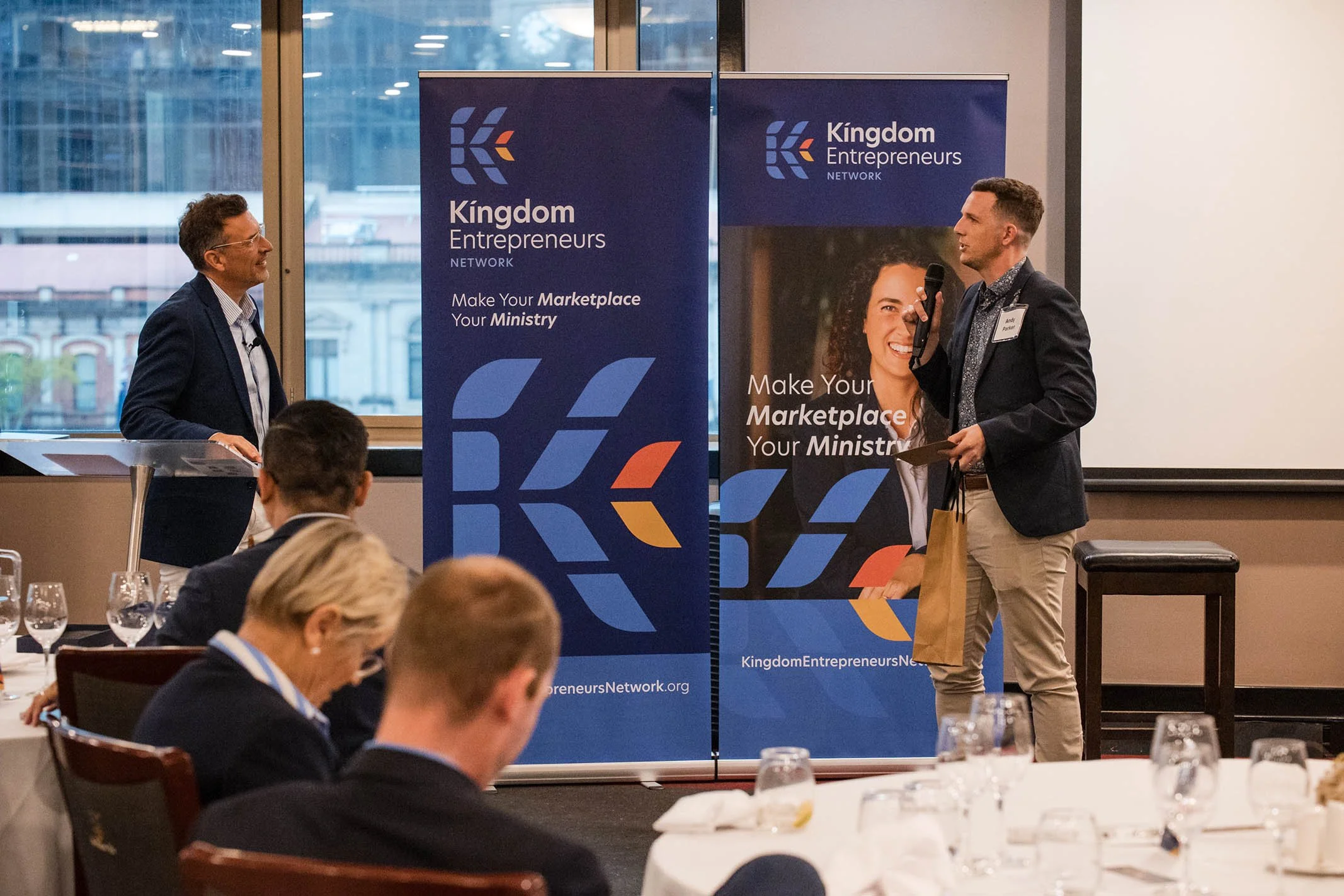A speaker and an interviewer are on stage at a conference, with banners from Kingdom Entrepreneurs Network behind them. Attendees are seated at round tables, taking notes.