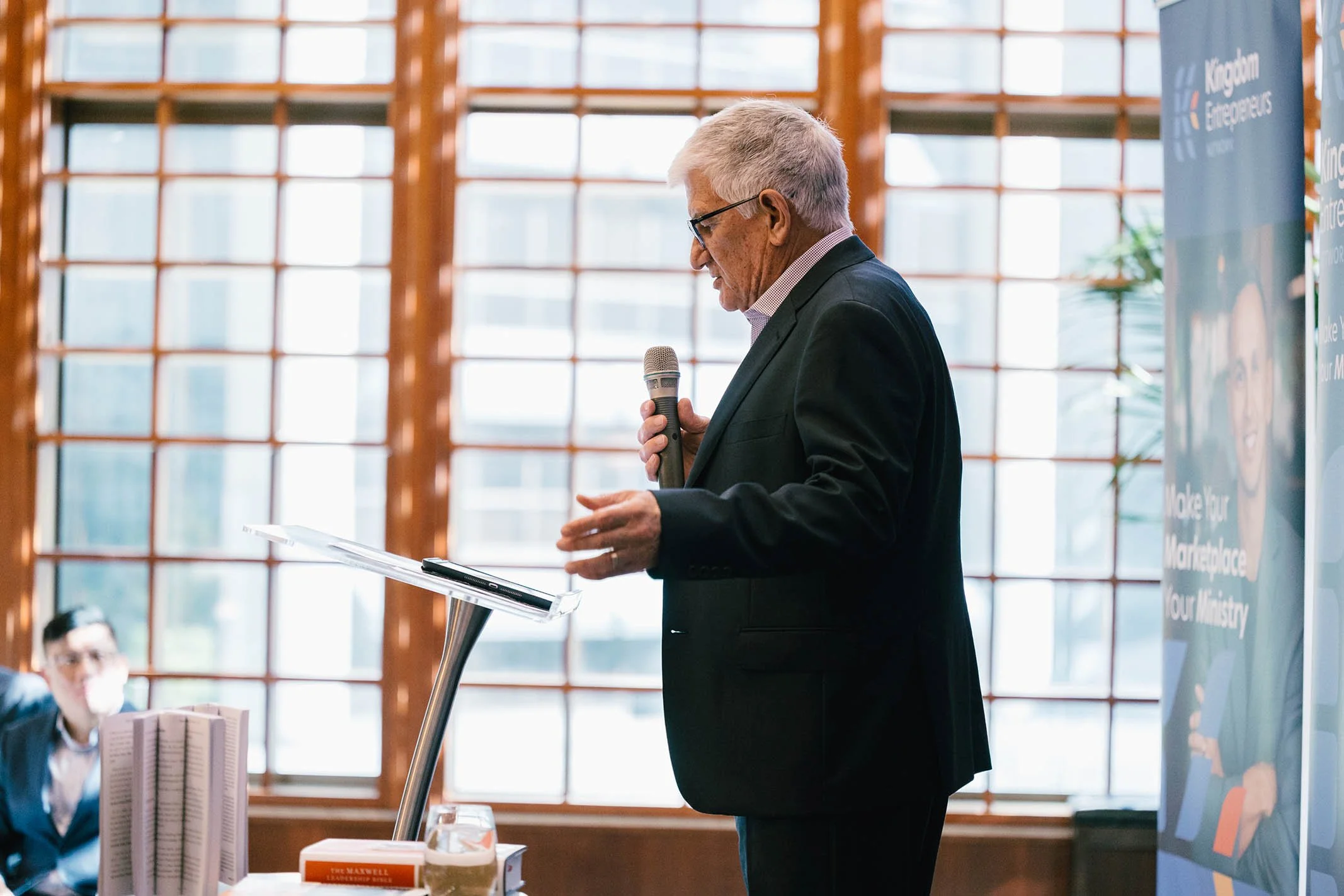 An elderly man in a black suit speaking into a microphone while looking at notes on a stand, with a large window in the background.